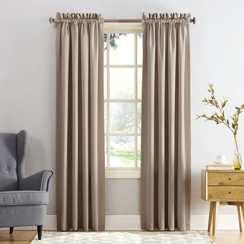 Beige rod pocket curtains hang over a window flanked by a gray wingback armchair and a wooden side table with two drawers.