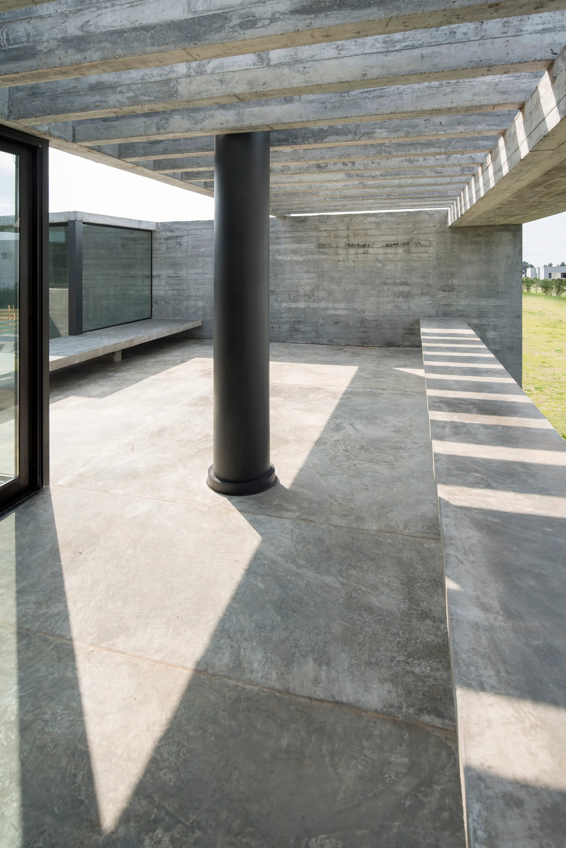 An outdoor concrete patio area featuring a central dark column, an overhead slatted pergola casting shadows, and low built-in concrete benches.