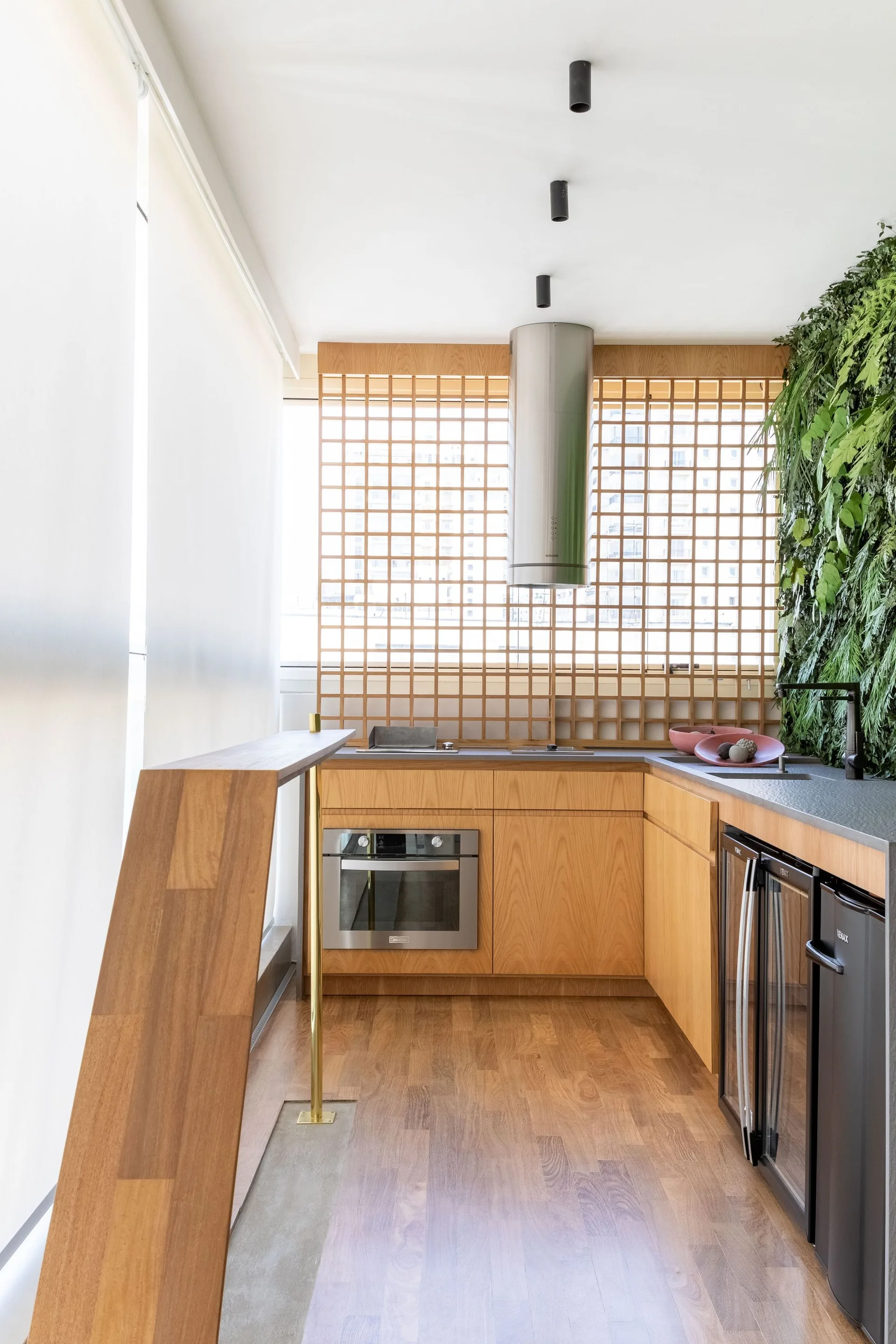 A modern wood-toned kitchen featuring a vertical green plant wall, an under-counter beverage fridge, and a wooden grid screen over the window.