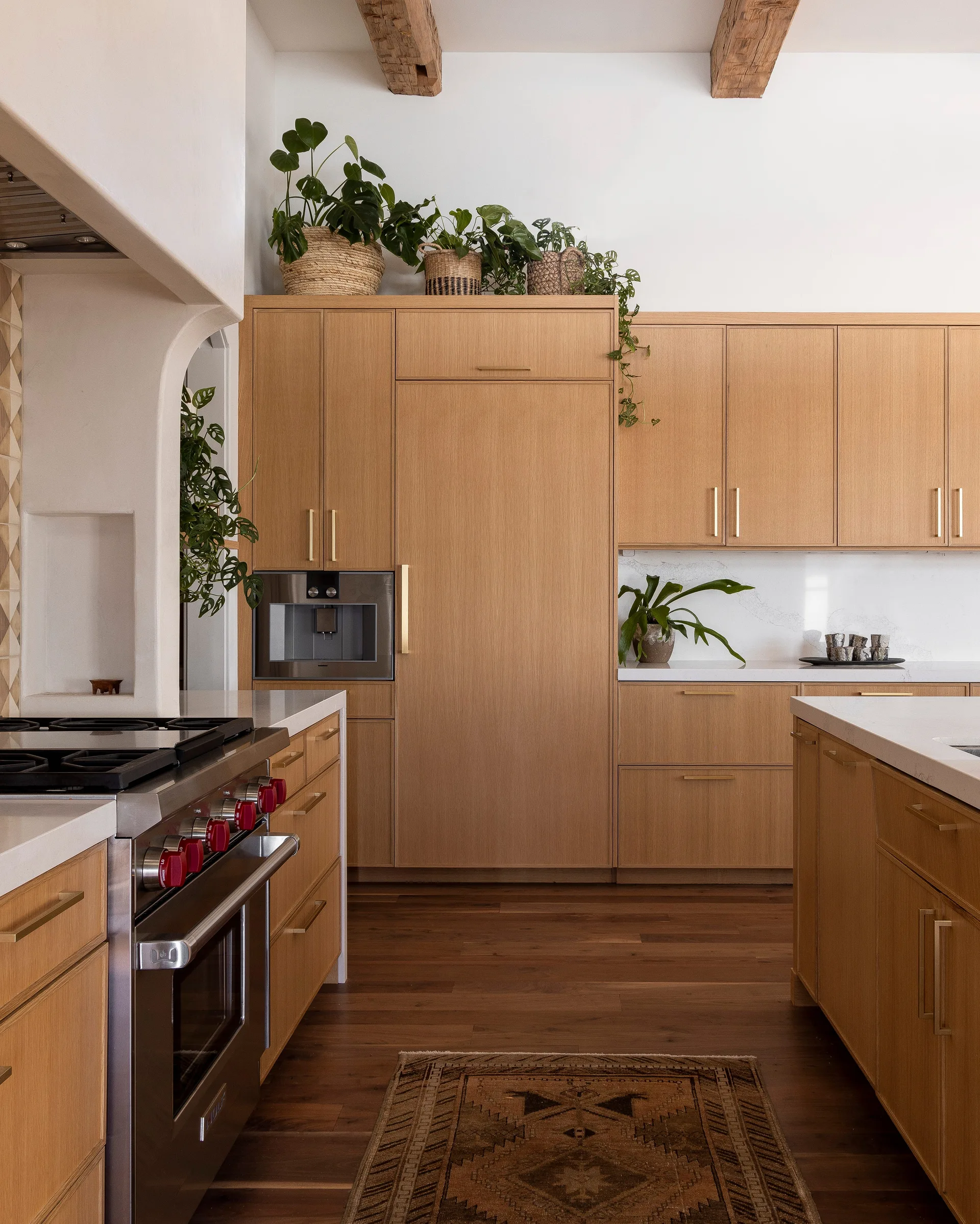 A modern kitchen featuring flat-panel light wood cabinetry, white stone countertops, a professional range with red knobs, and exposed ceiling beams.