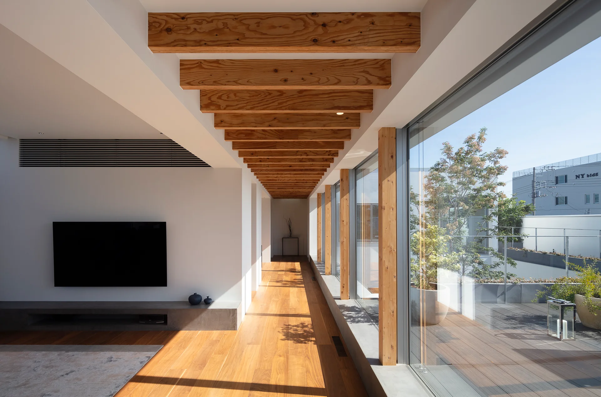 A modern living area with exposed timber ceiling beams, wood floors, a concrete media ledge beneath a wall-mounted TV, and large windows leading to a balcony.
