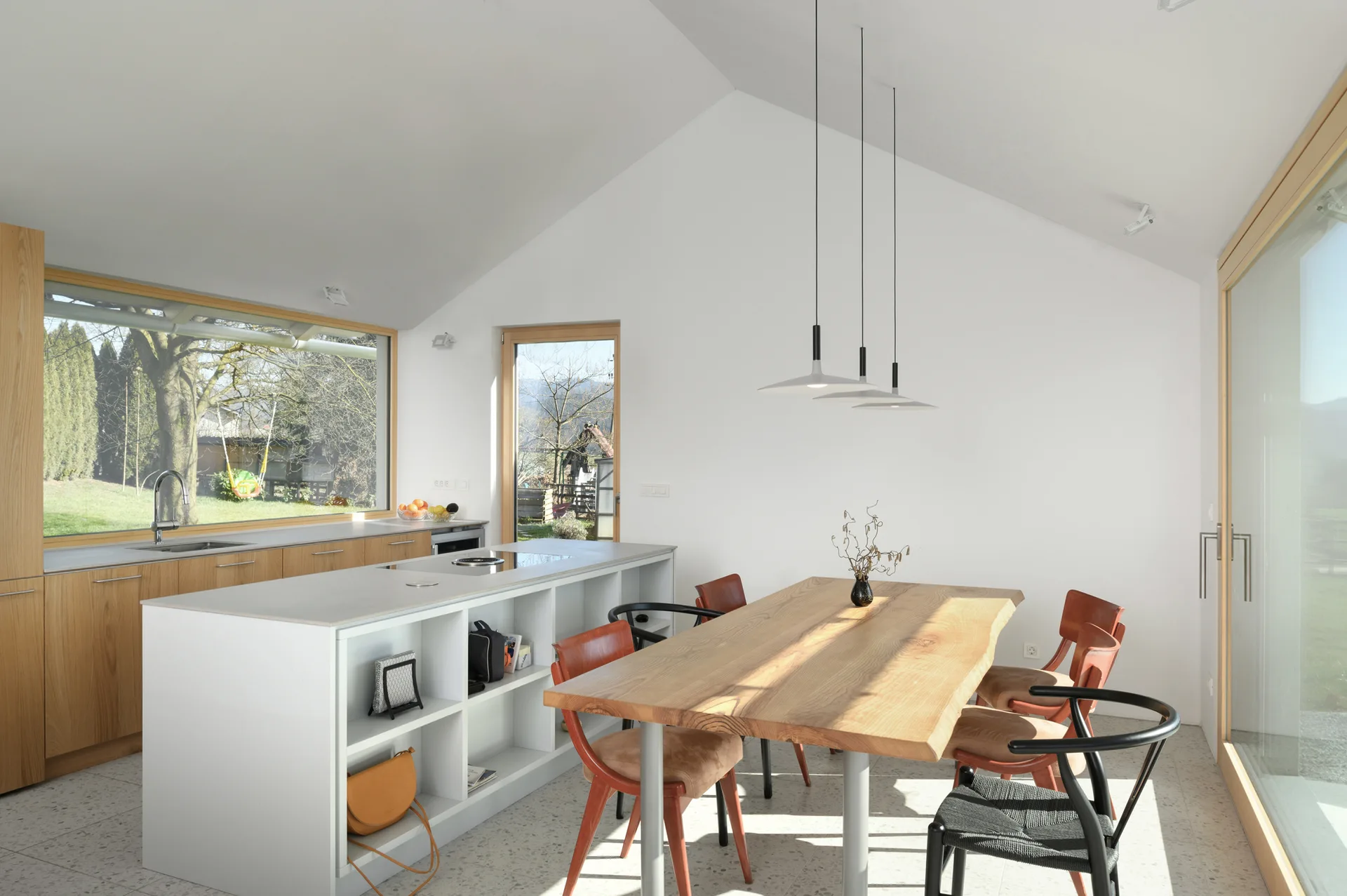 An open-plan kitchen and dining area featuring a vaulted ceiling, a white island, a live-edge wood table, and large wood-framed windows.