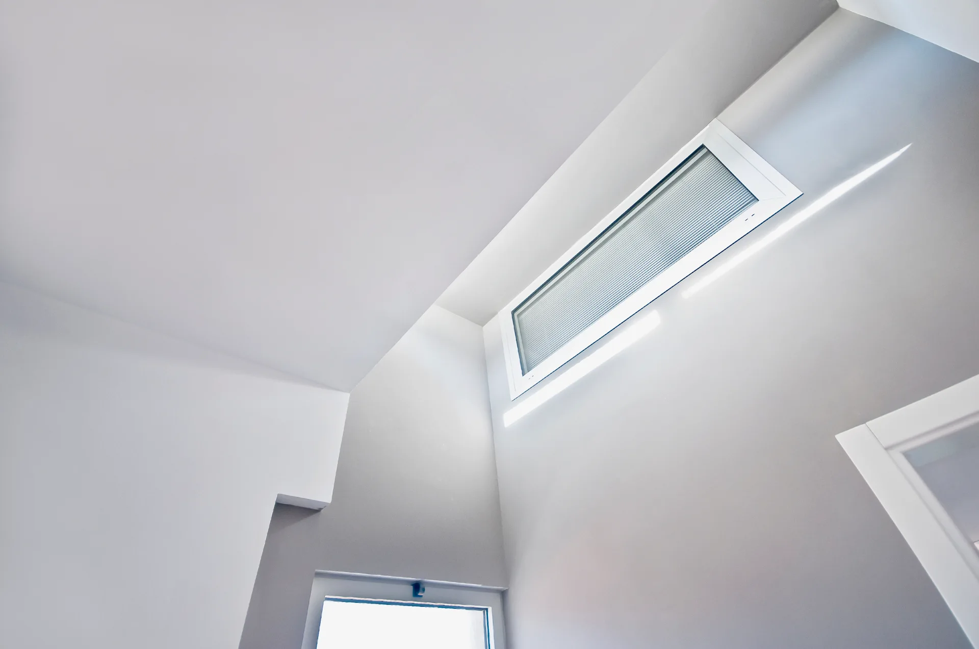 A minimalist interior corner featuring white painted walls, a sloped ceiling, and a high-set rectangular window.