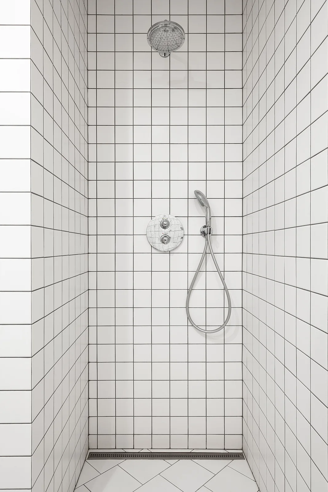 A minimalist shower stall features white square wall tiles in a grid pattern, polished chrome fixtures, and a linear floor drain.