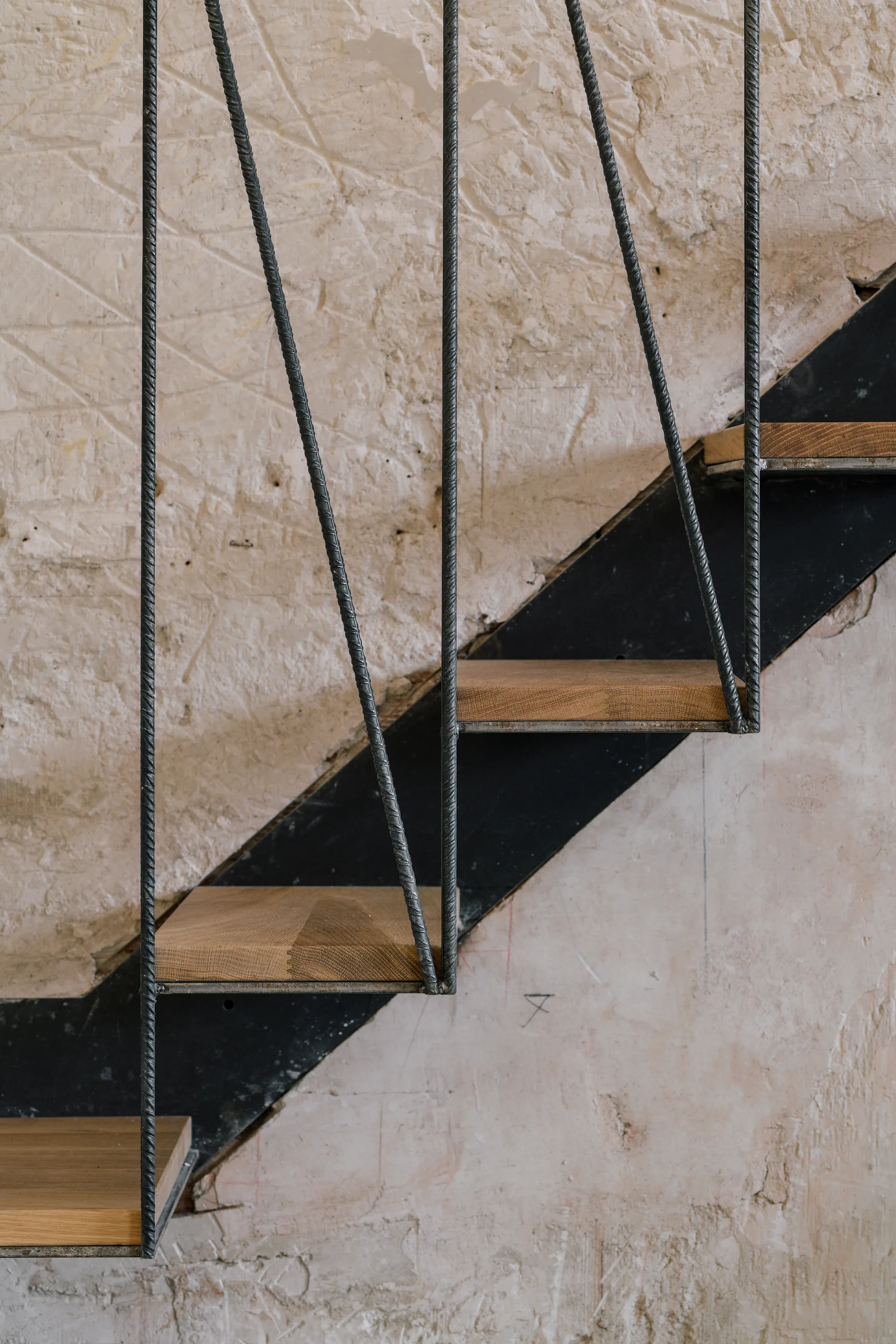 Close-up of an open-riser staircase with wooden treads, vertical metal rebar balusters, and a black steel frame against an unfinished plaster wall.