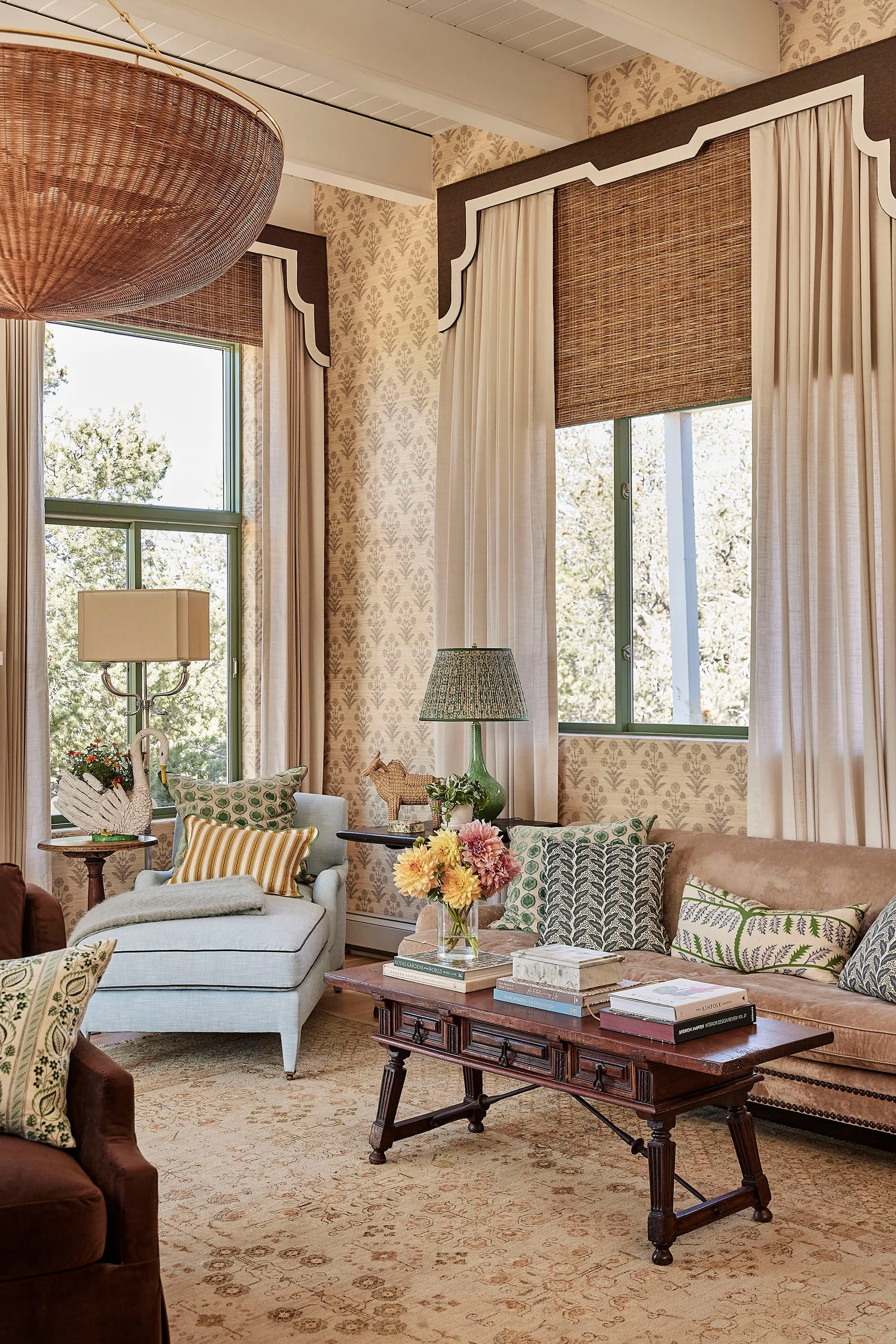 A traditional living room with a tan sofa, light blue chaise lounge, botanical wallpaper, and windows with woven wood shades and drapery.