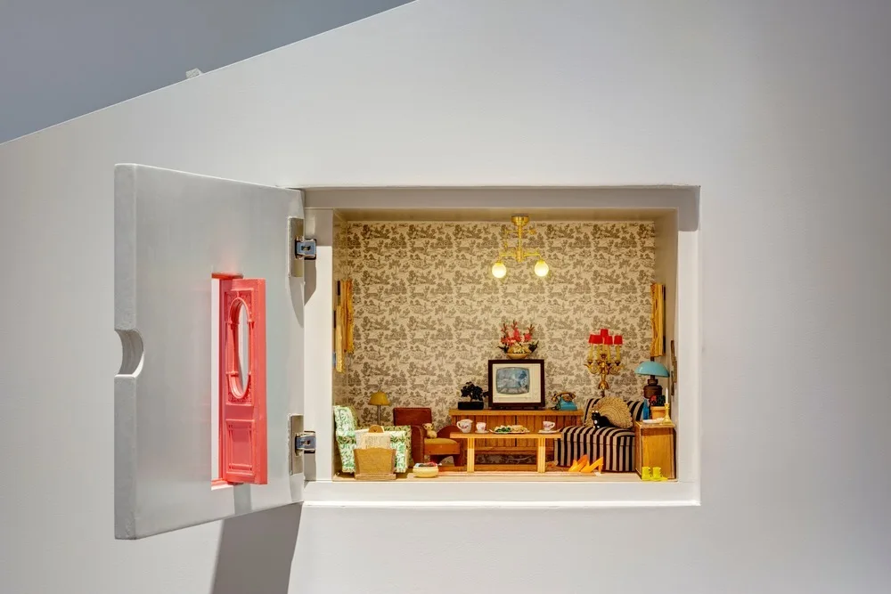 A miniature living room display built into a white wall niche, featuring tiny retro furniture, striped upholstery, and toile wallpaper.