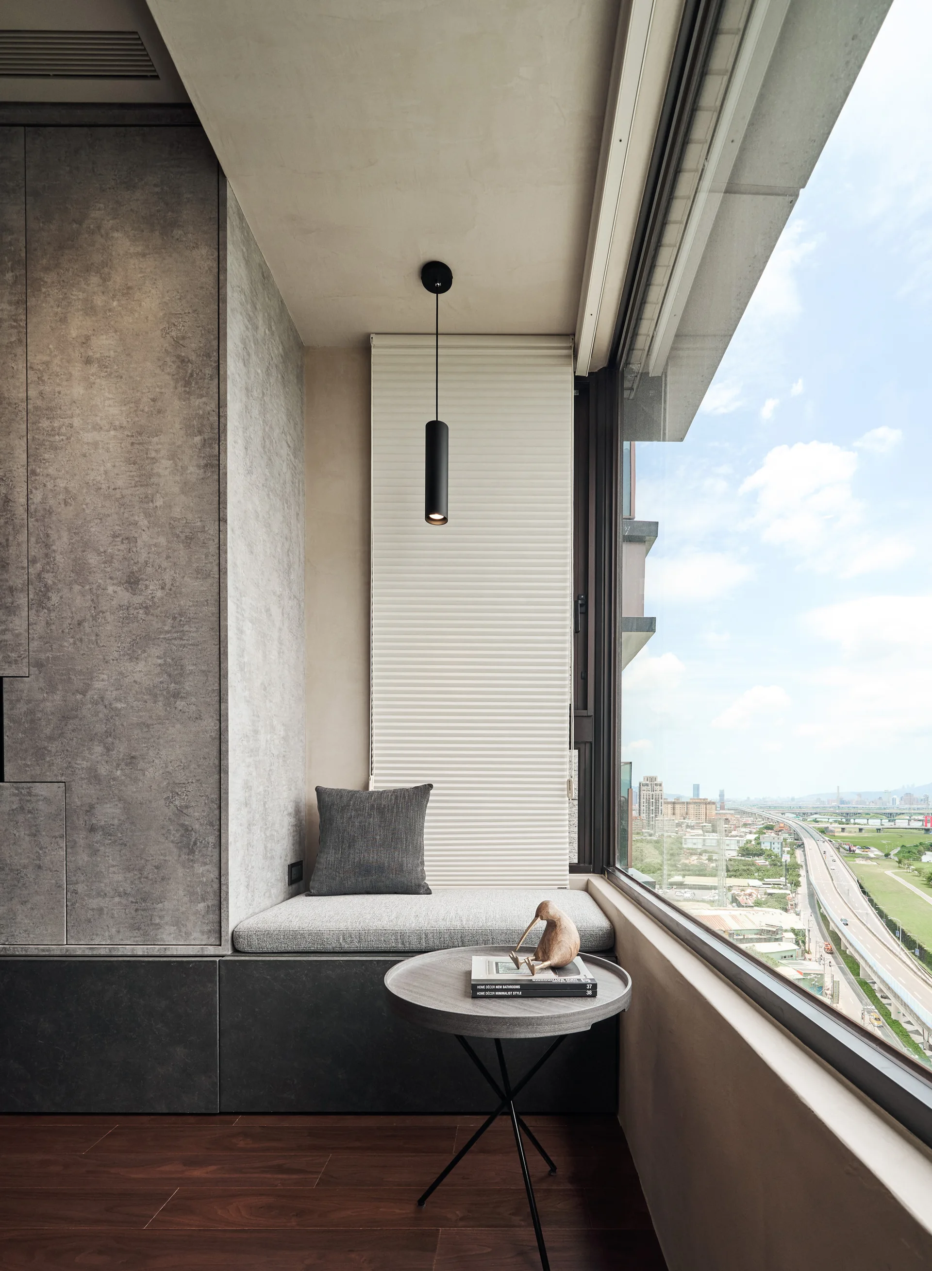 A modern built-in window seat features a gray cushion, a black pendant light, and a small round side table next to a large window overlooking a city.