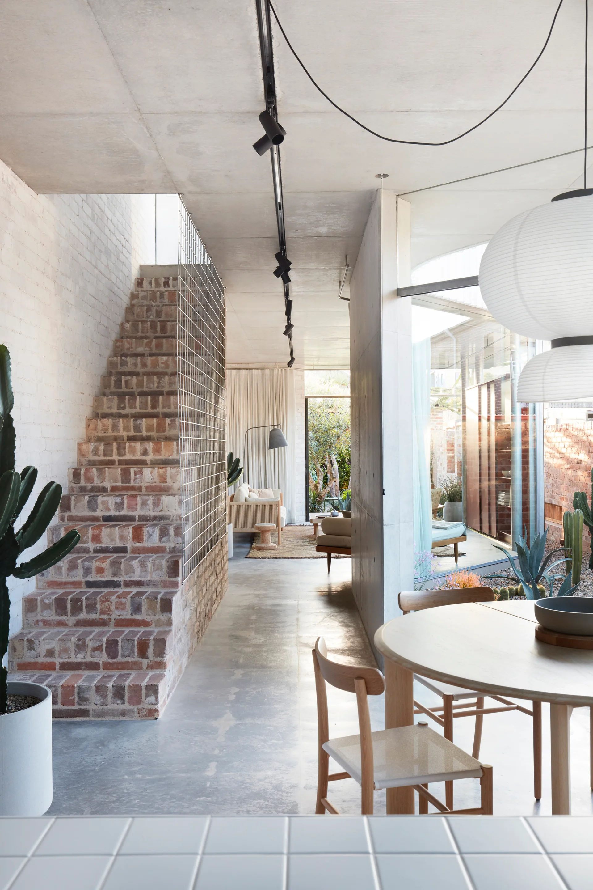 A modern home interior with an exposed brick staircase, concrete floor and ceiling, wooden dining table, and large glass windows.