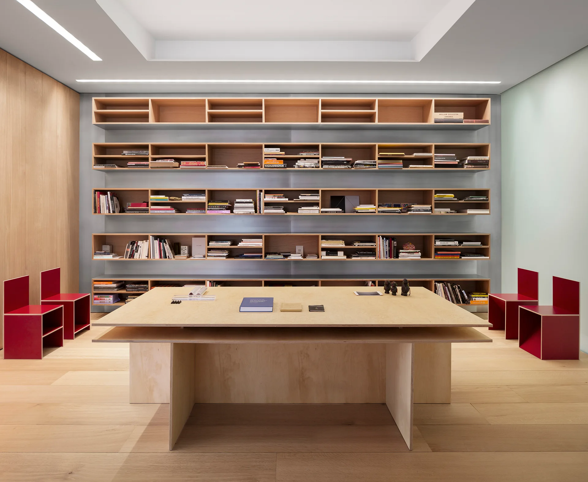 A minimalist workspace features a large central plywood table, wall-to-wall wooden bookshelves, and four red block-style chairs on light wood floors.