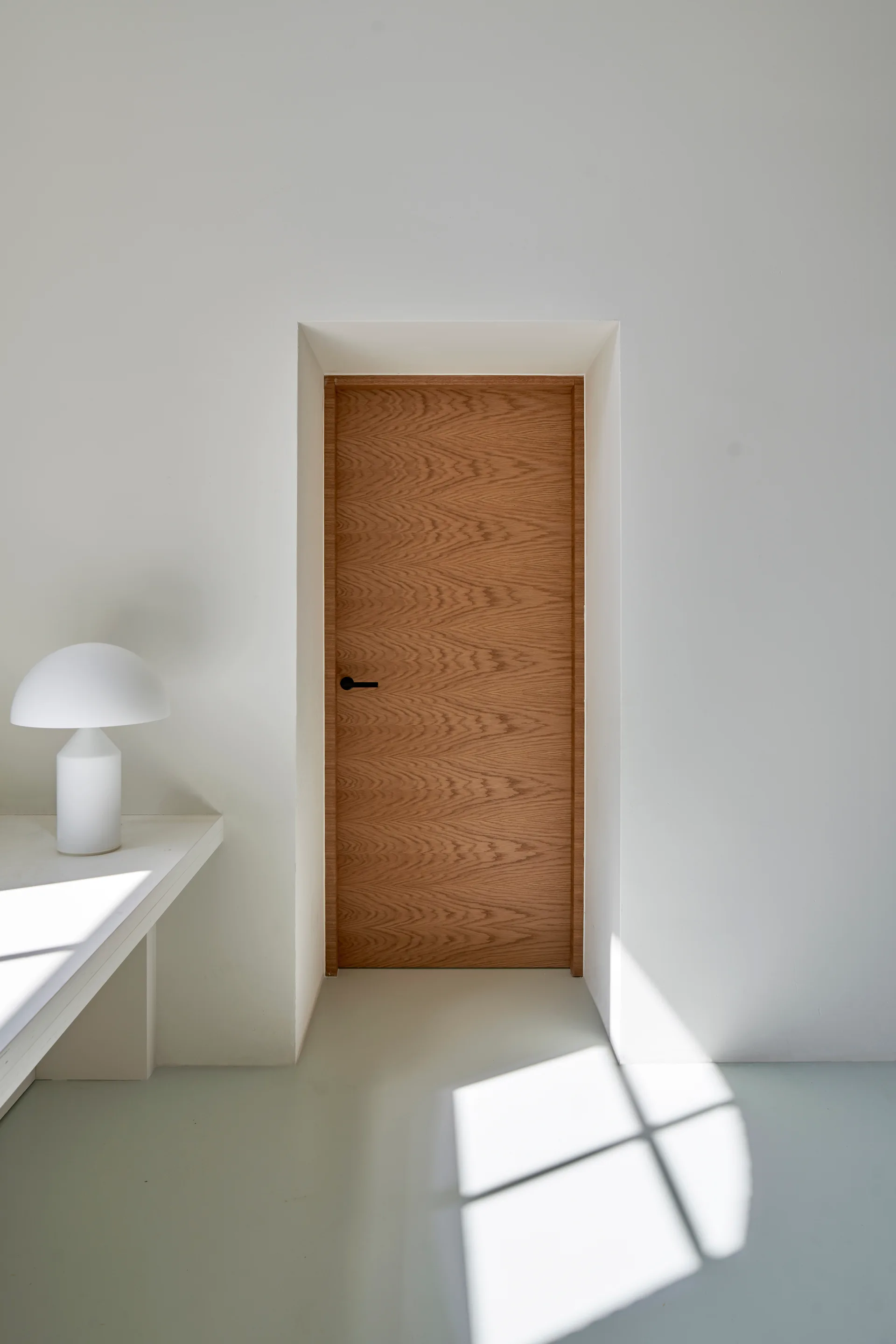 A wooden door with a black handle is recessed into a plain white wall next to a white mushroom lamp on a console.