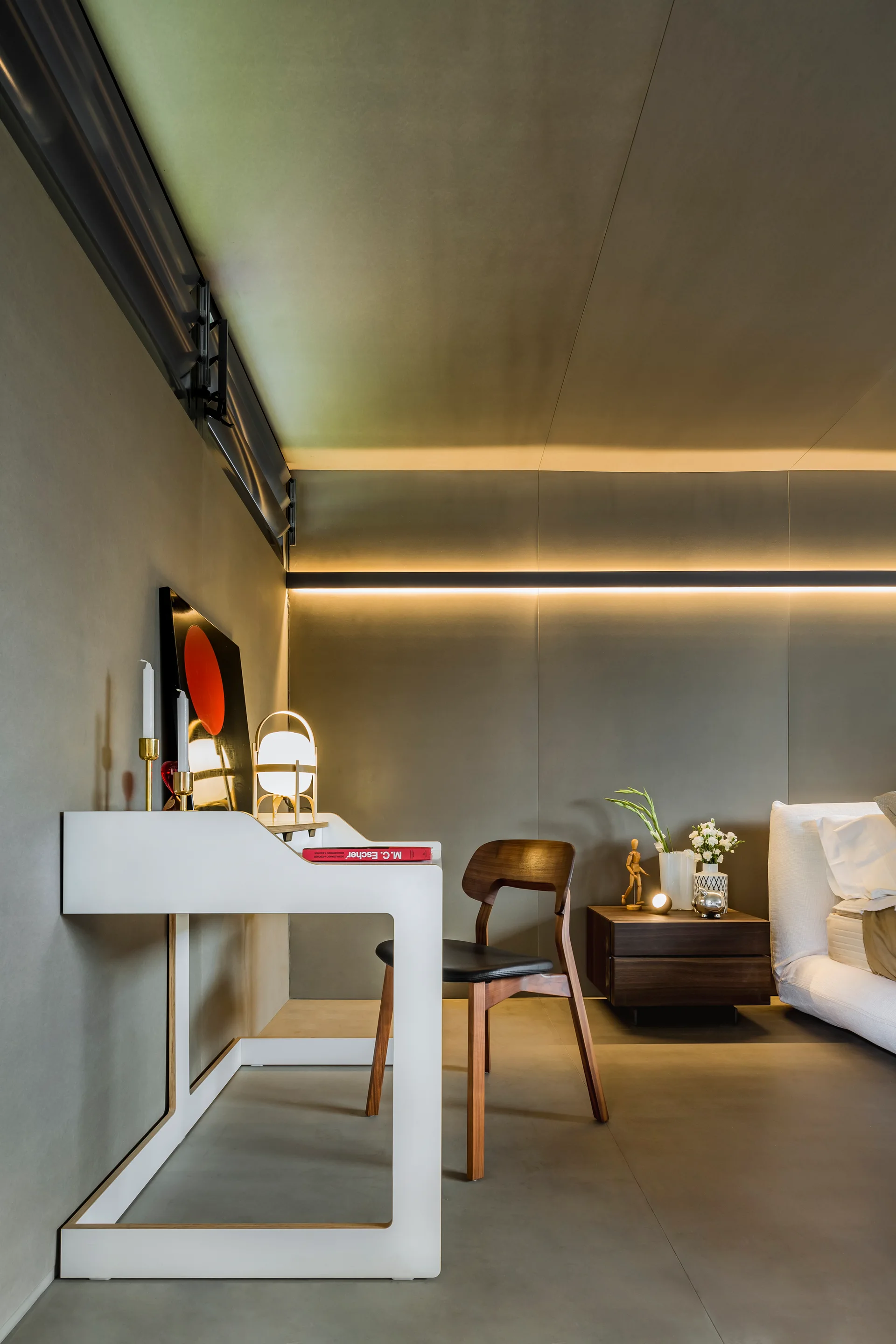 A minimalist bedroom features grey paneled walls, an illuminated horizontal light strip, a white writing desk with a wooden chair, and a low nightstand.