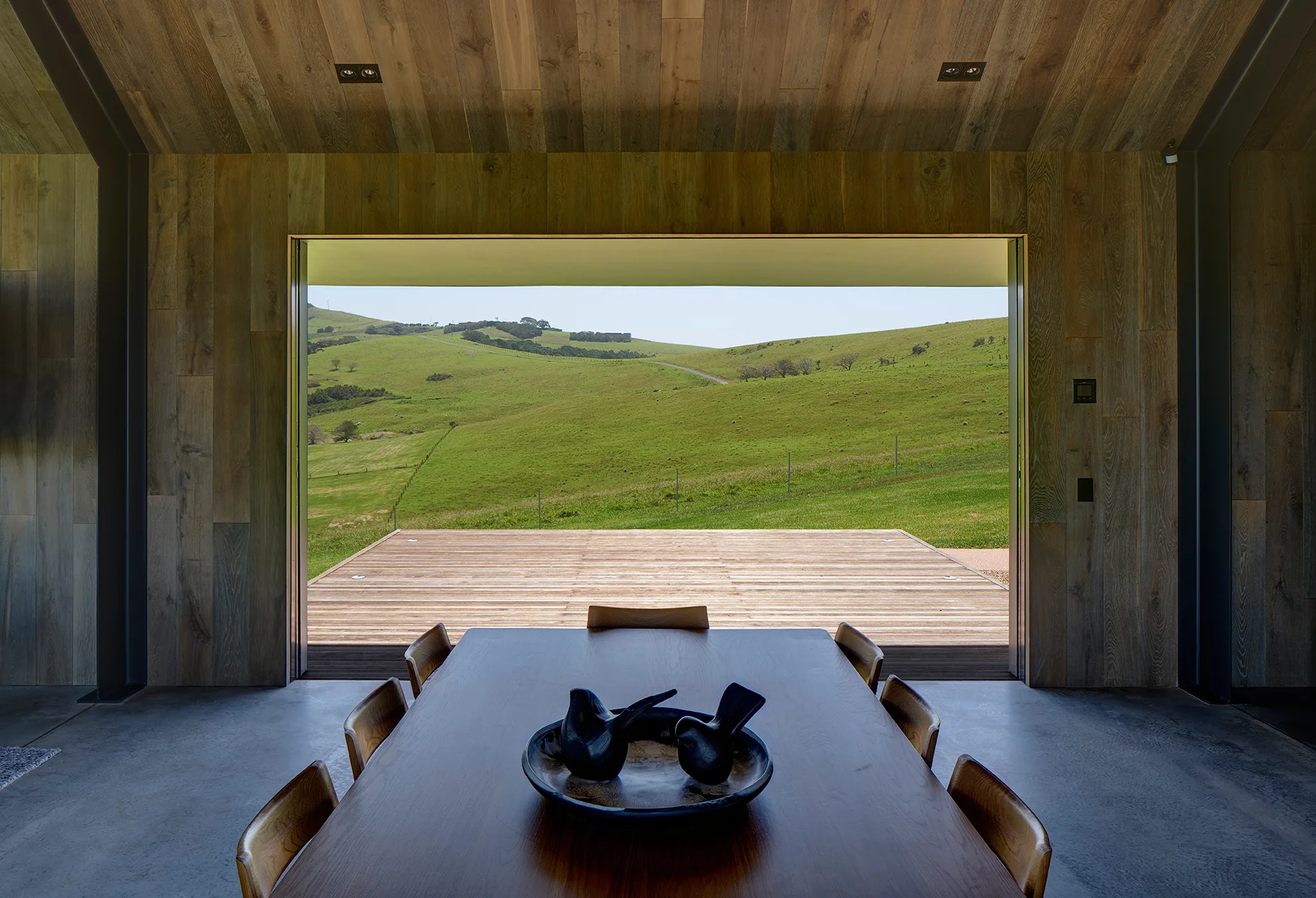 A minimalist dining room clad in wood paneling features a long wooden dining table and chairs facing a wide open view of rolling green hills.