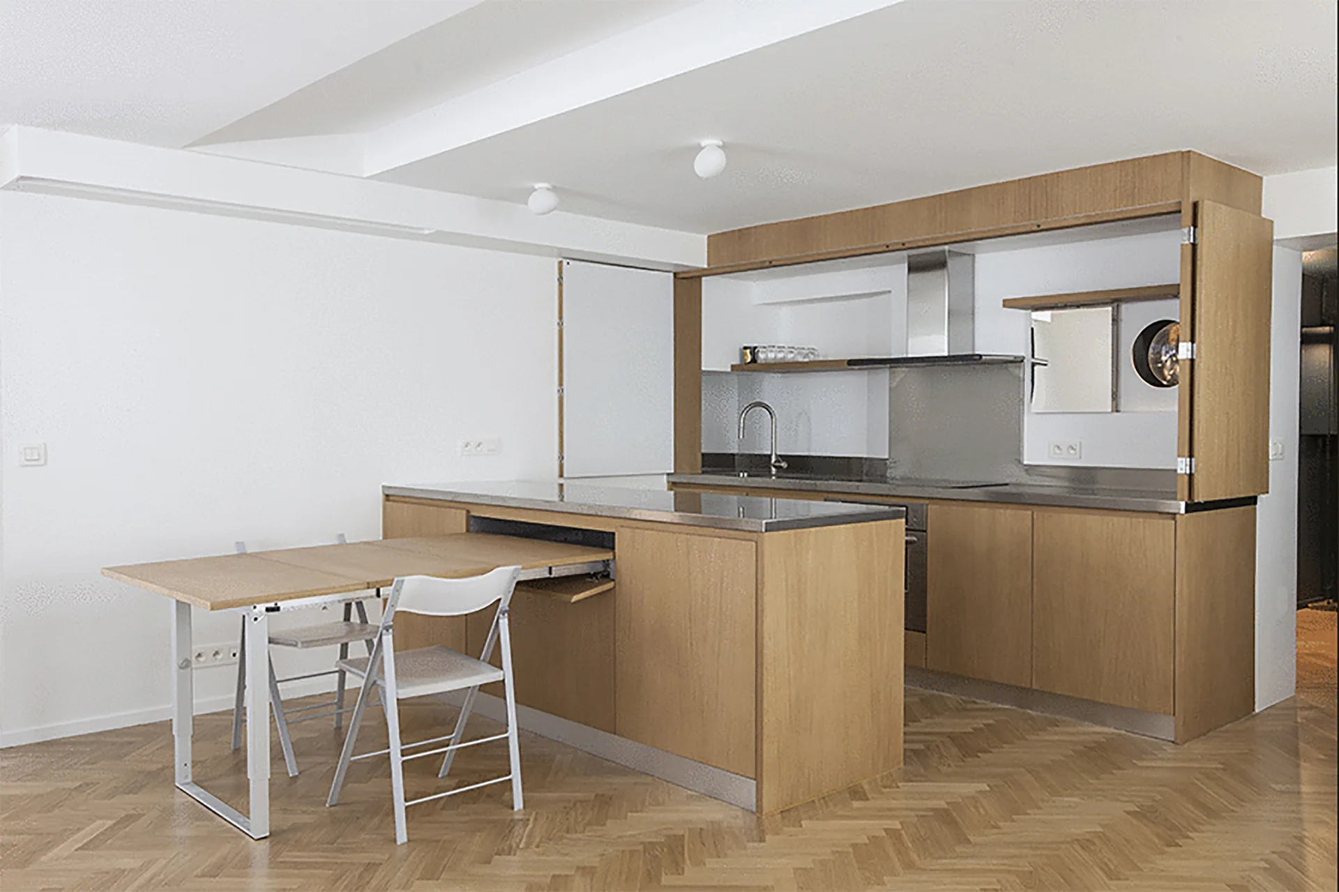 A minimalist kitchen features a light wood island with an extended pull-out dining table, white folding chairs, and herringbone wood floors.
