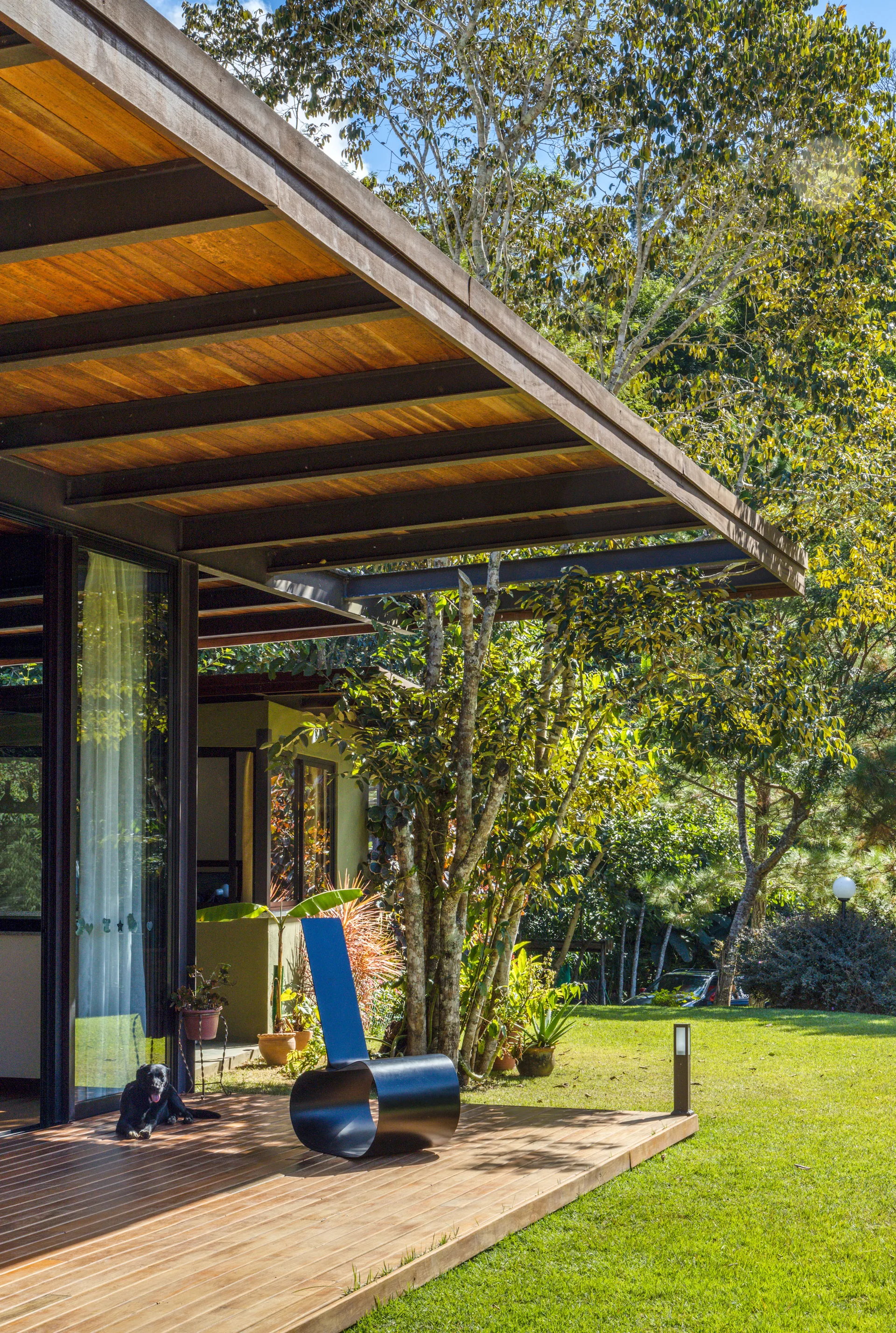 A modern wooden deck features a sculptural curved black lounge chair under an architectural wood overhang overlooking a grassy garden.
