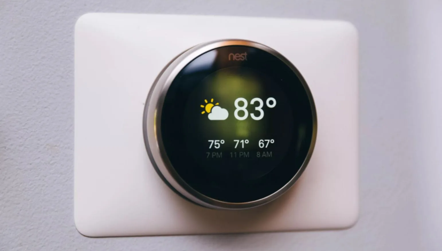 A close-up of a circular smart thermostat with a black screen and silver ring, mounted on a white rectangular plate against a light gray wall.