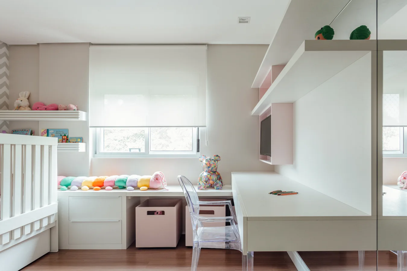 A bright children's room features a white wooden crib, an integrated desk with a clear acrylic chair, and soft pink storage boxes.