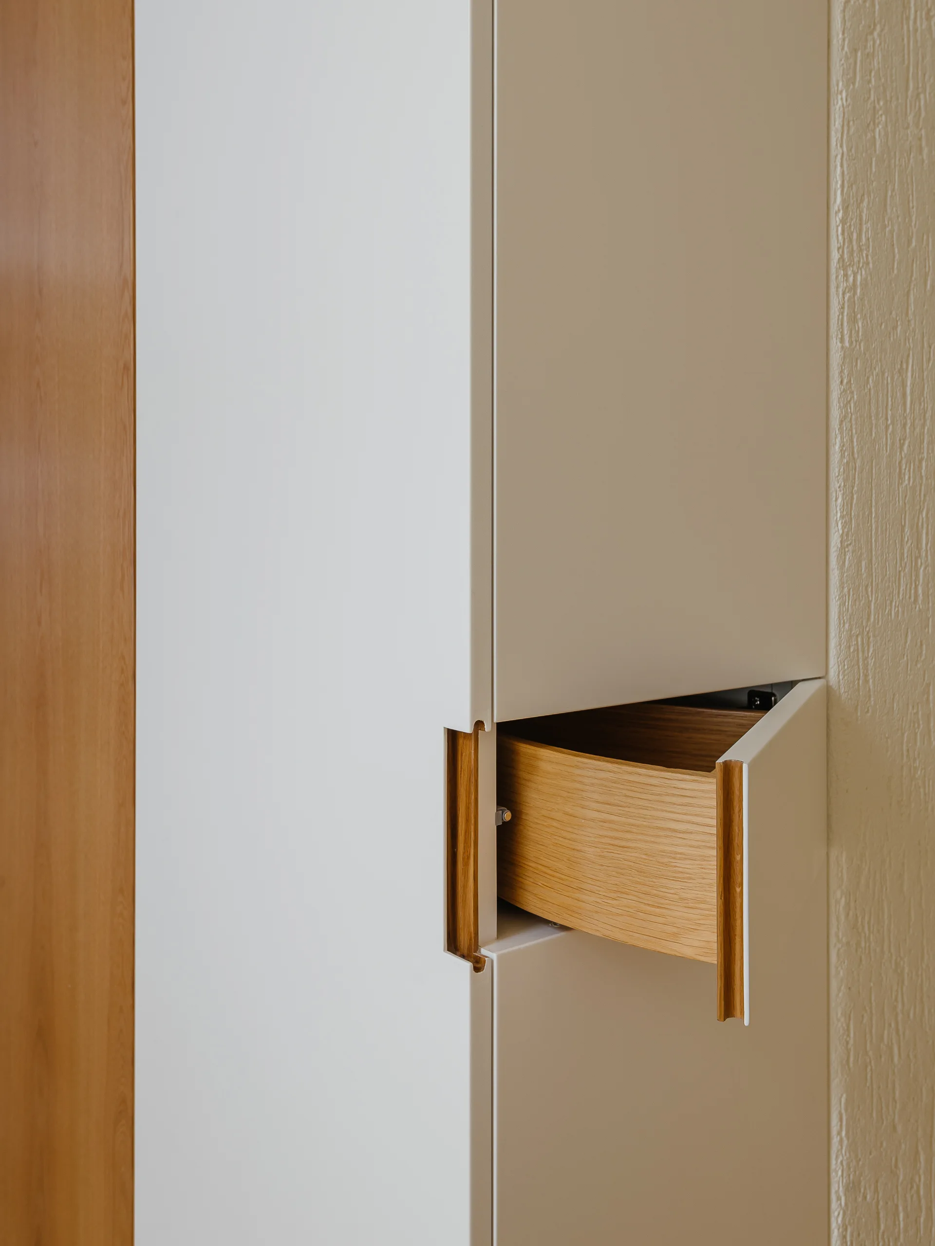 A close-up of a minimalist cream cabinet featuring an integrated wooden recessed handle and a partially open wooden drawer.