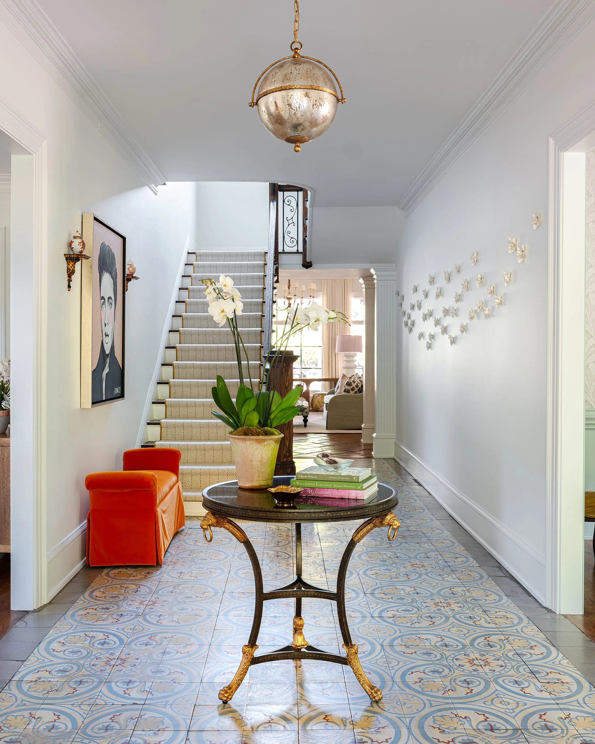 A bright residential foyer displays blue patterned tile flooring, a central round gold-trimmed table with an orchid, and a staircase with a runner.
