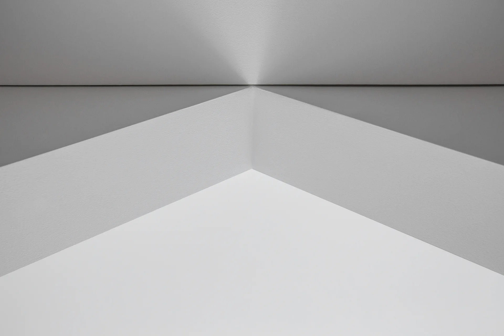 Minimalist interior wall corner showing a clean architectural junction with sharp geometric angles and directional shadows in light grey and white.