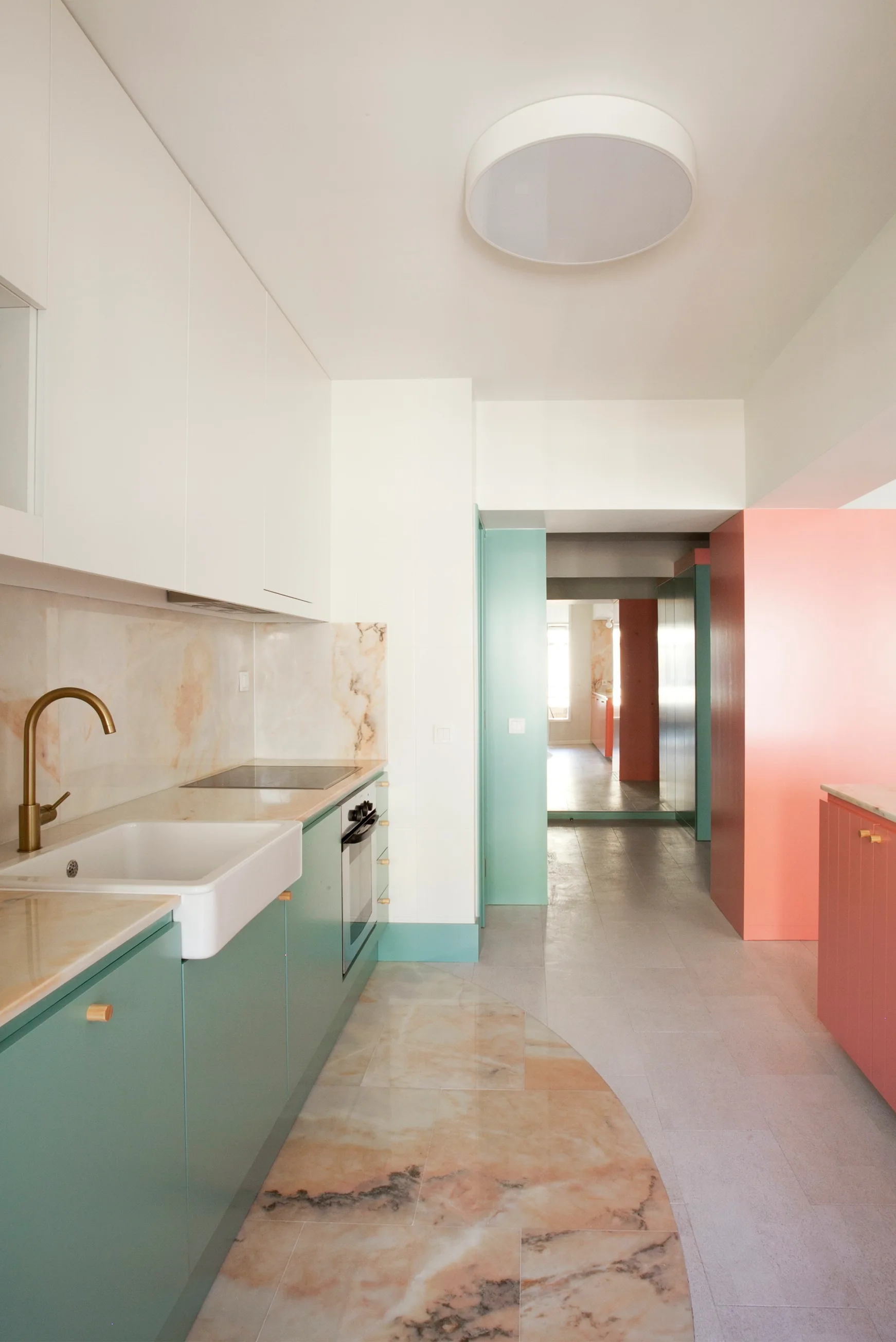 A modern kitchen featuring matte teal lower cabinets, white upper cabinets, coral painted walls, a brass faucet, and a semi-circular marble floor inlay.