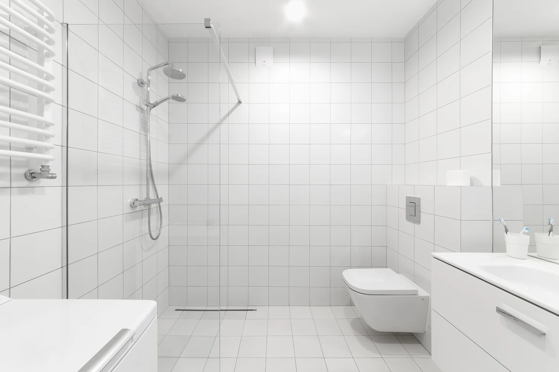 A minimalist white bathroom featuring walls and floors covered in square tiles, a glass shower panel, and a wall-mounted toilet.