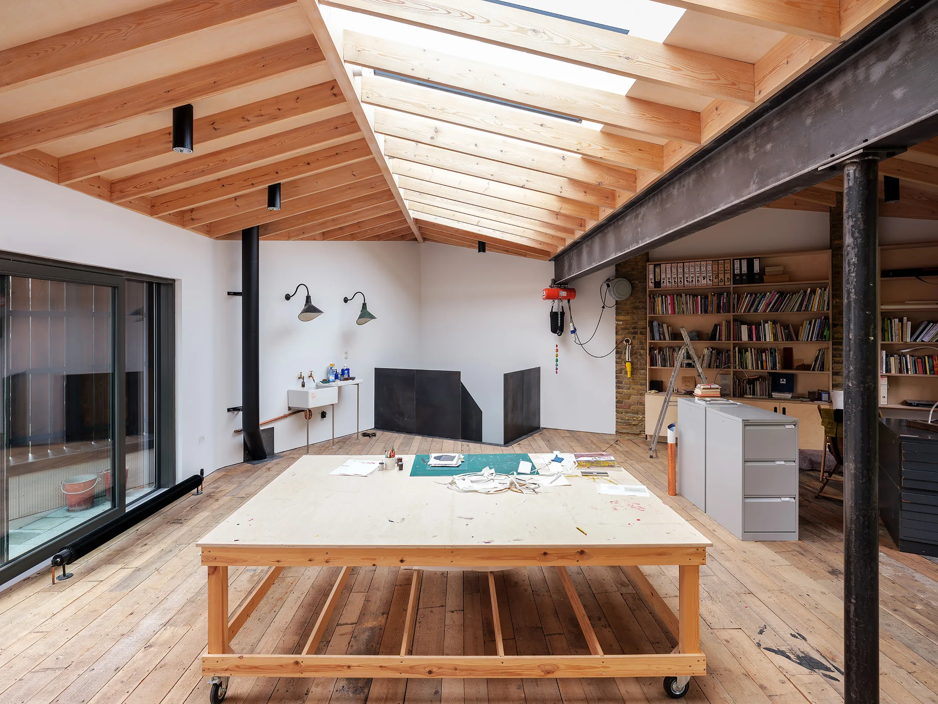 An artist studio features a large wooden work table on casters beneath a vaulted ceiling with exposed timber beams, skylights, and a steel support beam.