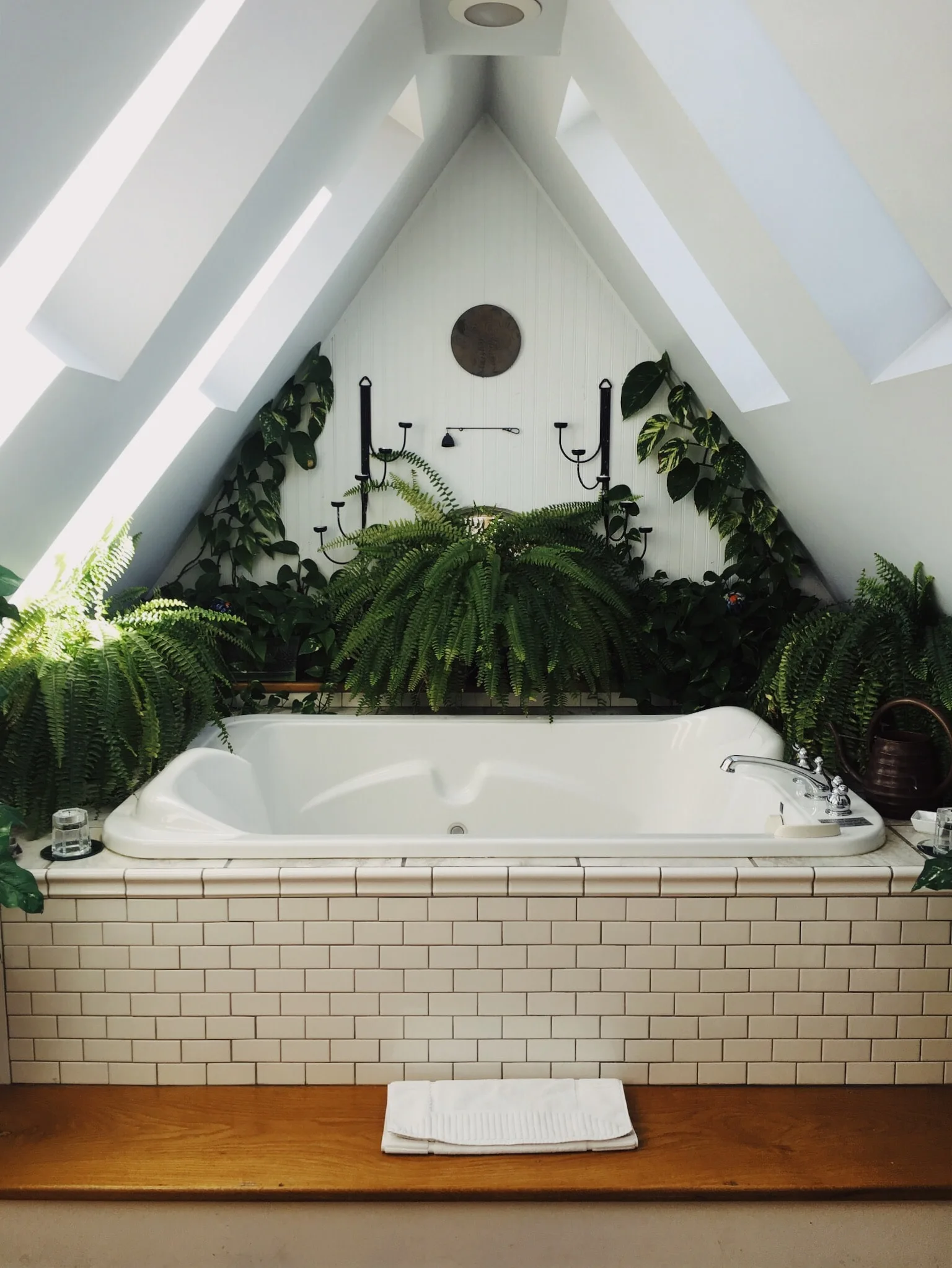 A white soaking tub with subway tile surrounds sits in an attic alcove beneath two skylights and overflowing green plants.