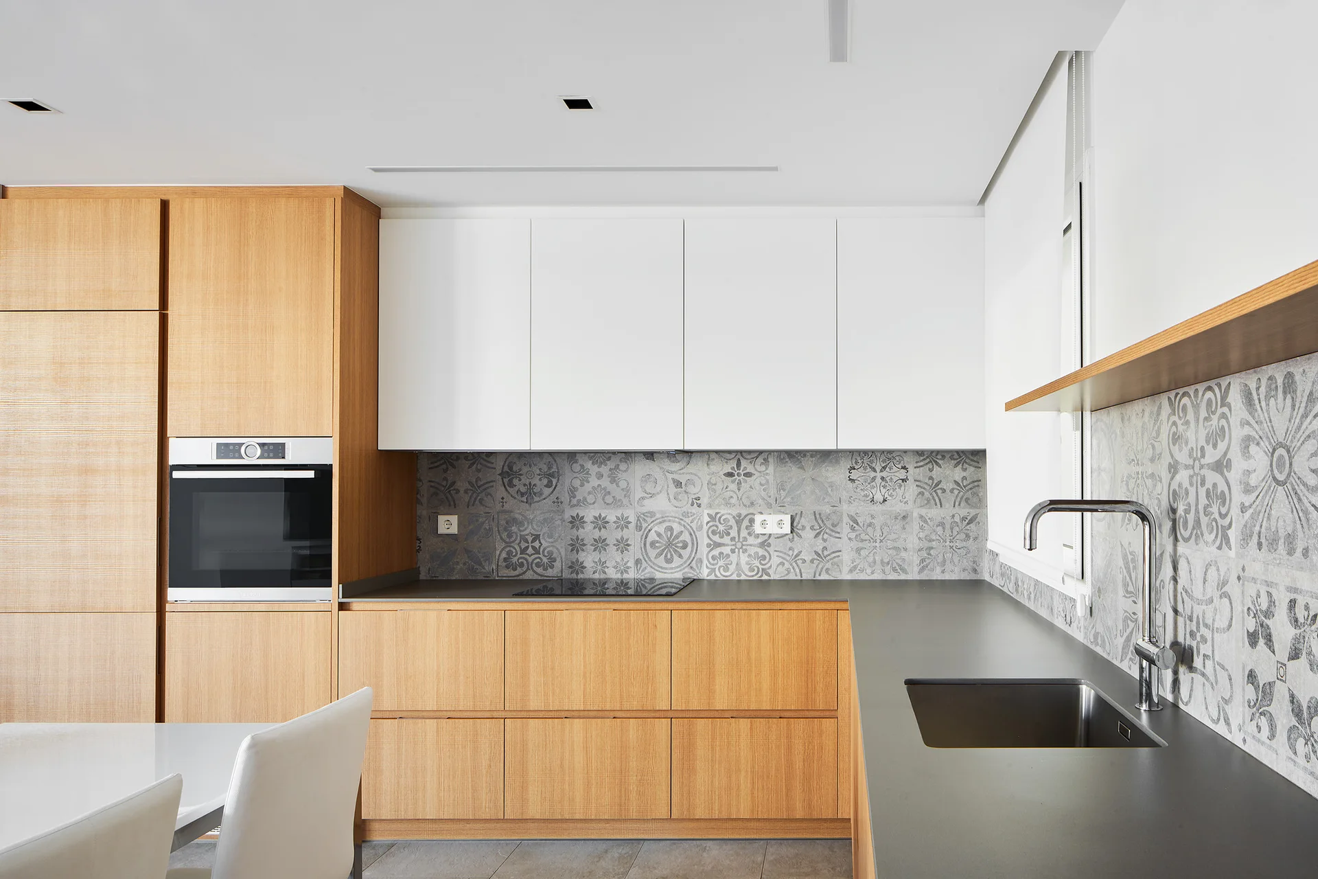 A modern kitchen features light wood lower cabinets, white upper cabinets, a dark grey countertop, and a patterned grey tile backsplash.