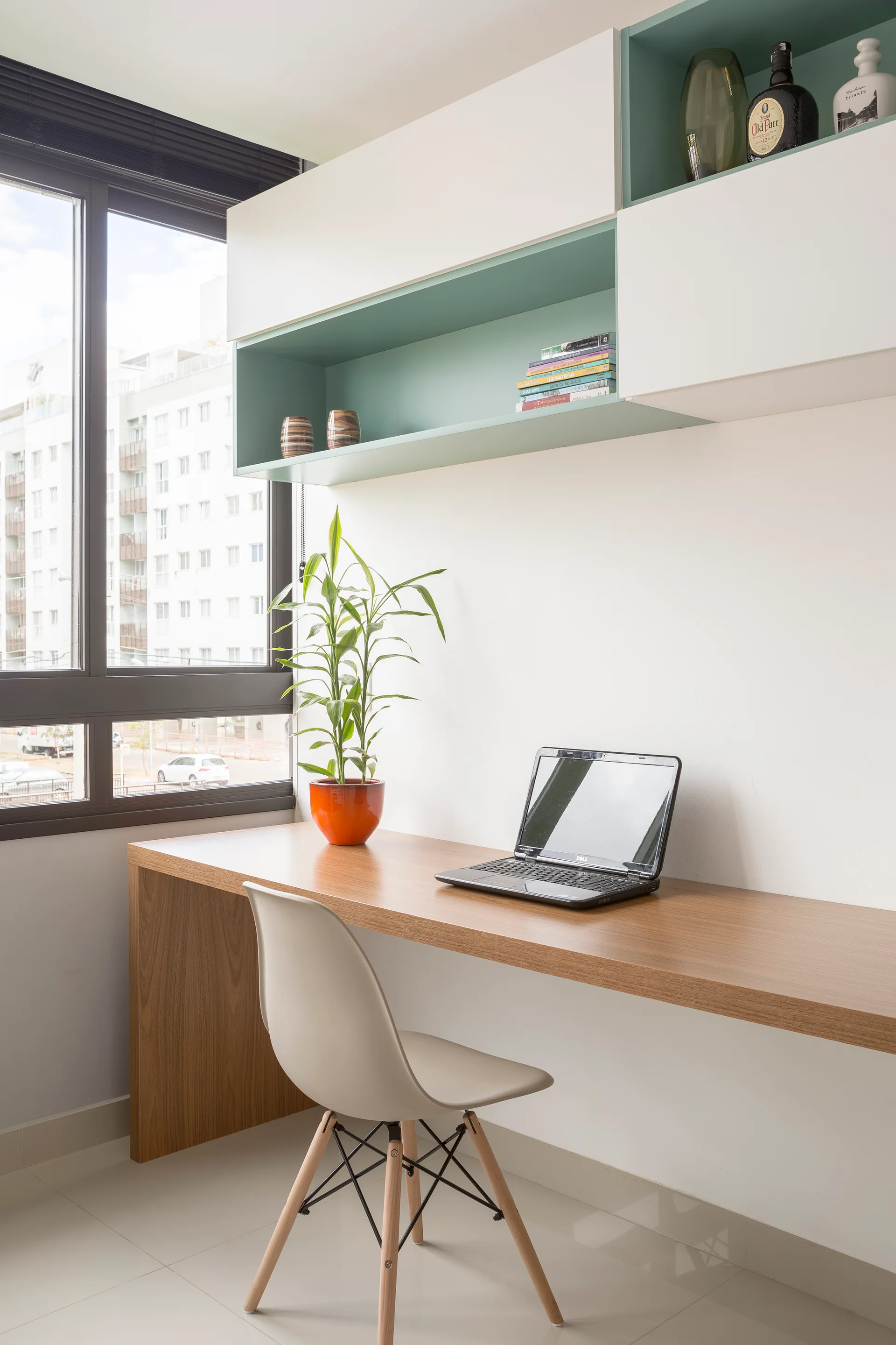 A bright home office workspace features a long wood desk, a molded chair, and white and mint green floating shelves next to a large window.