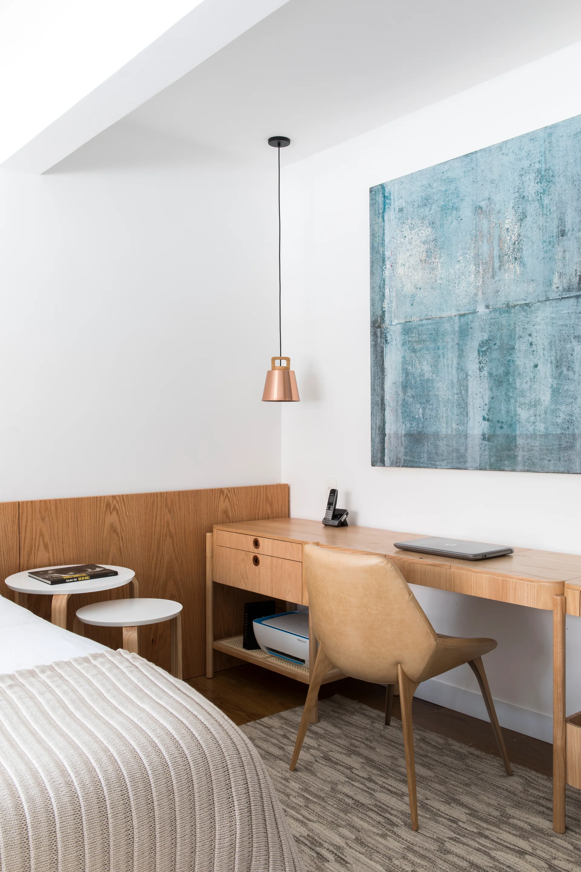A bedroom workspace features a light wood desk, a tan leather chair, a hanging copper pendant light, and a large abstract blue painting on the wall.