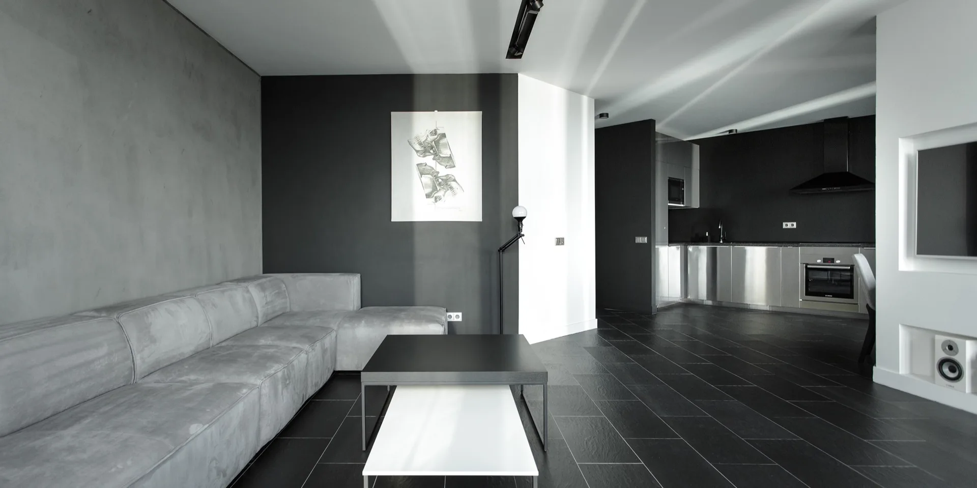A minimalist living room features a grey sectional sofa, nested black and white coffee tables, and black slate flooring with a kitchen in the background.
