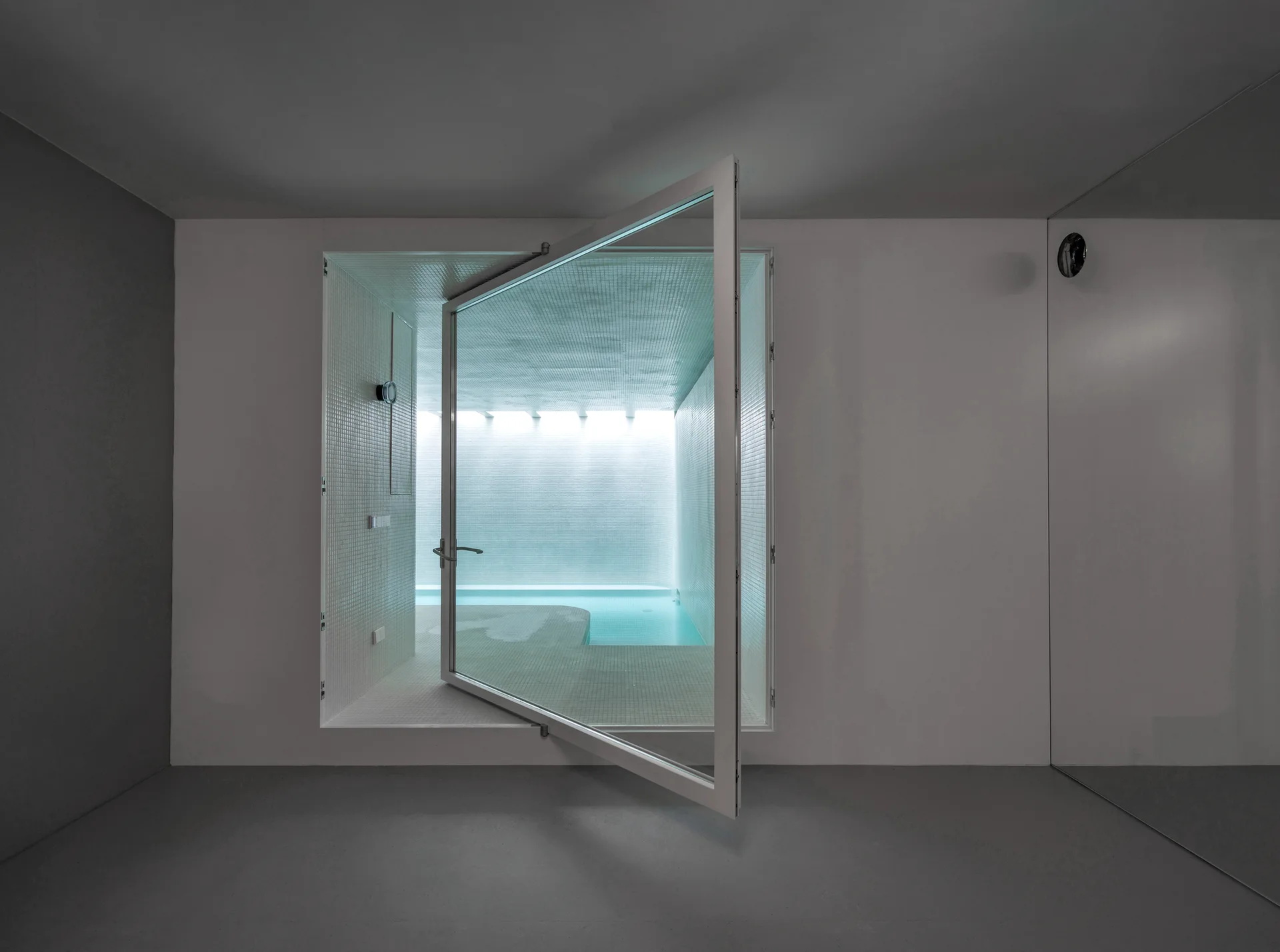 A large white-framed glass pivot door opens from a smooth grey room into a bright, white-tiled indoor pool area with glowing blue water.