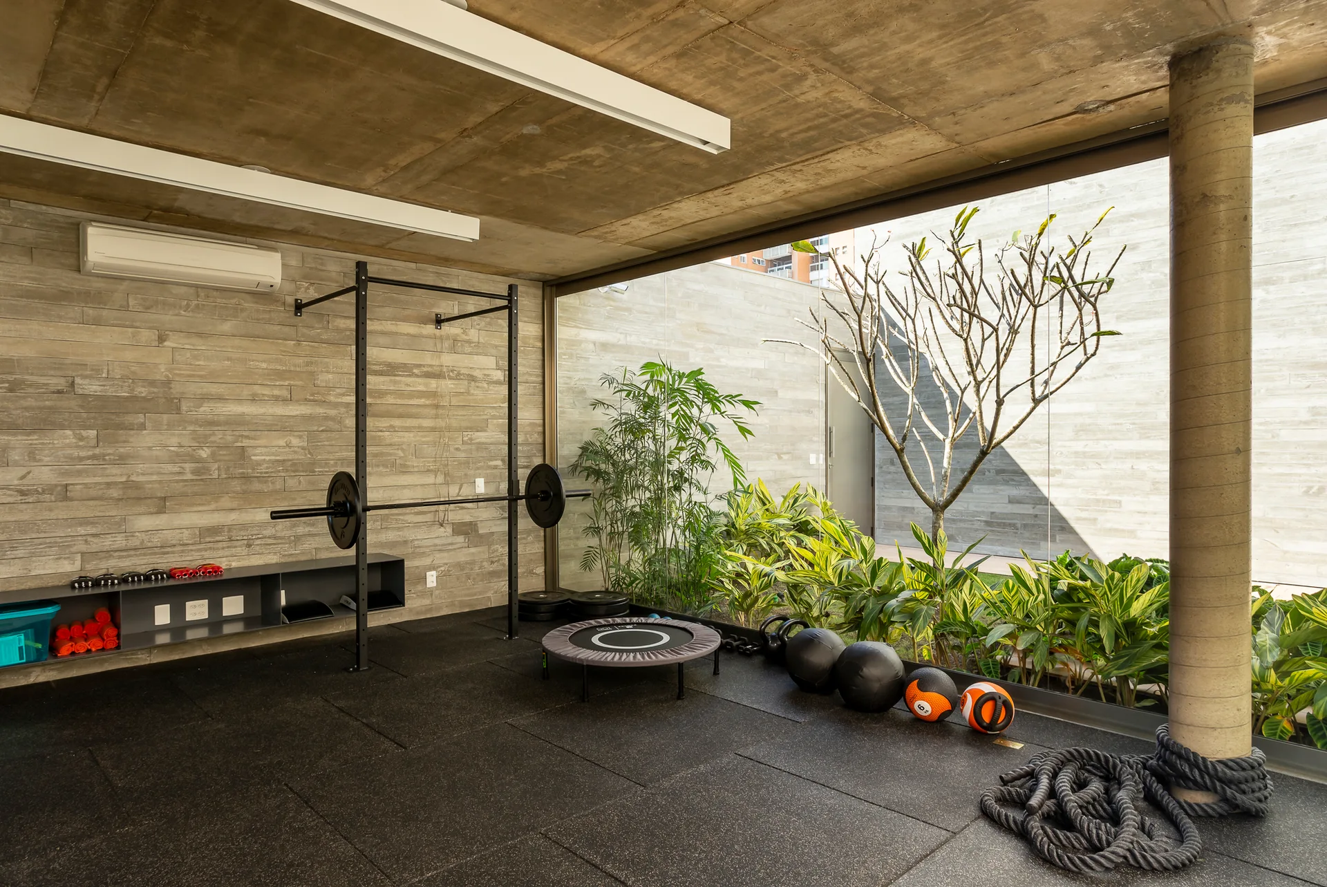 An industrial-style home gym featuring an exposed concrete ceiling, black rubber floor mats, and a large glass wall looking out to a garden.