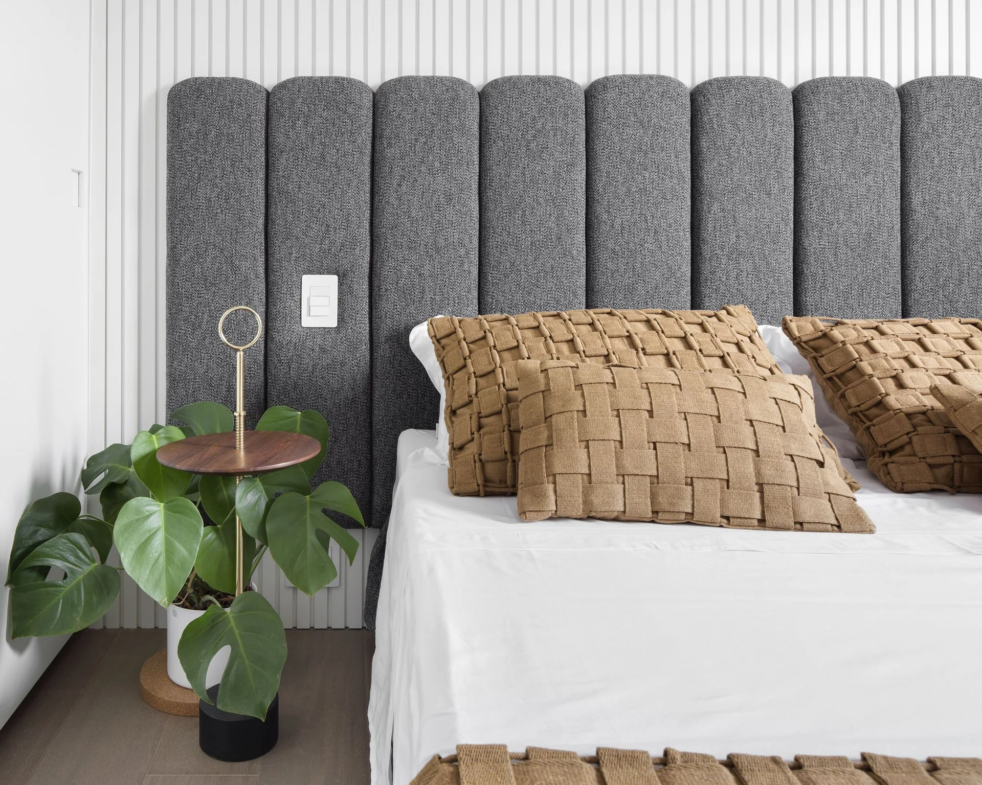 A grey upholstered panel headboard stands against a white vertical slat wall, flanked by woven tan pillows on the bed and a leafy monstera plant.