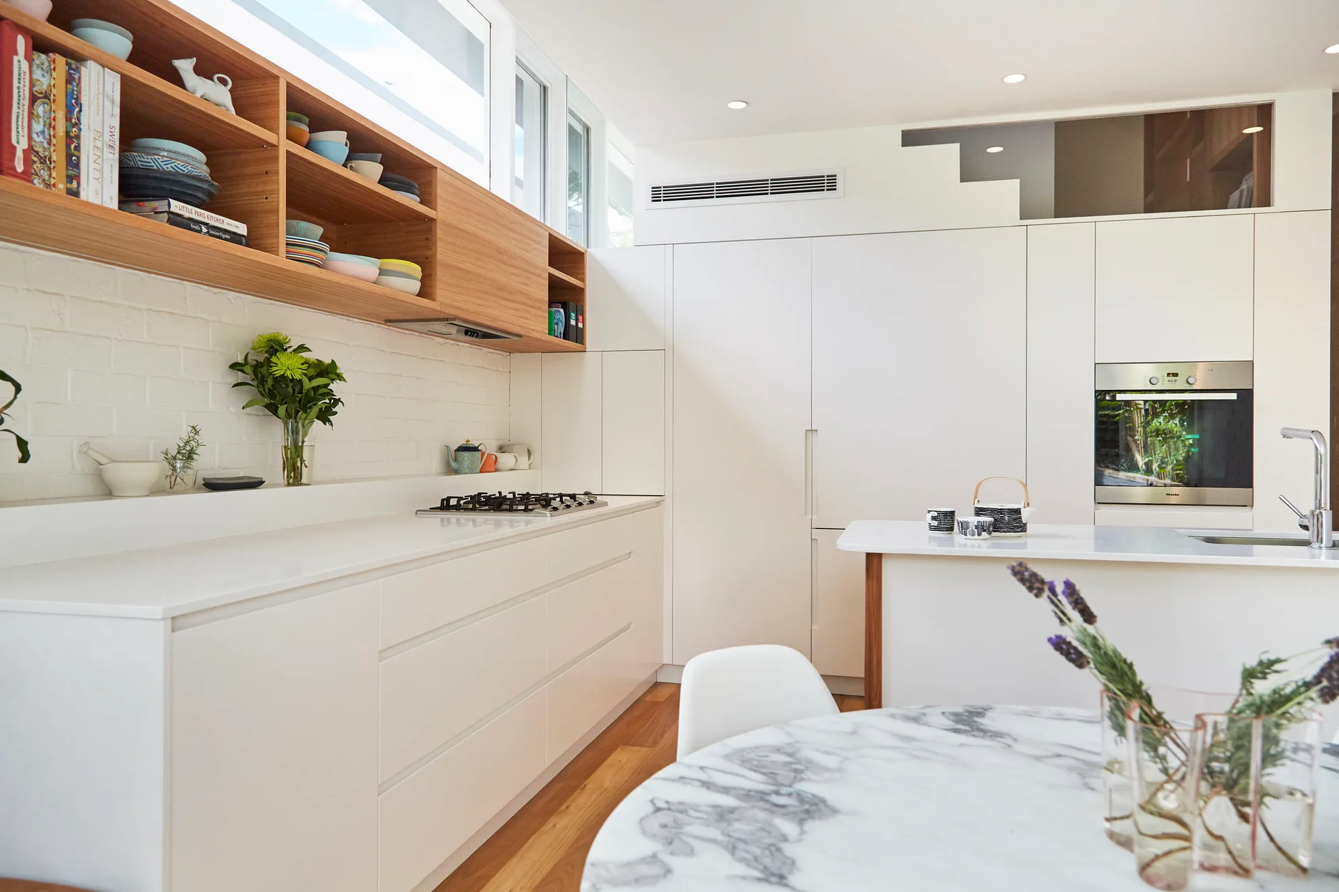 A modern white kitchen featuring flat-front cabinetry, wood open shelving below clerestory windows, and a round marble-topped dining table.