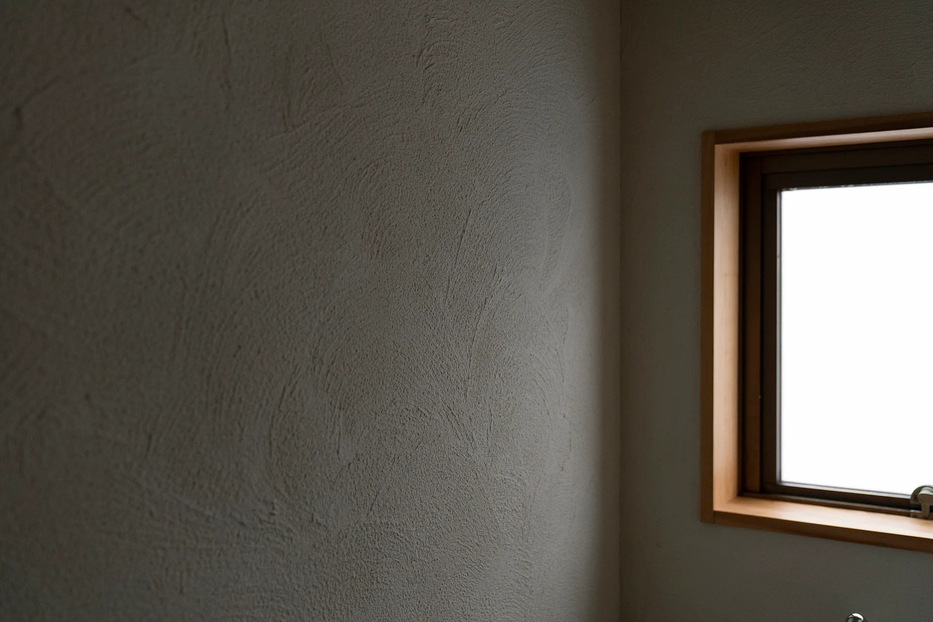 A close-up of a hand-troweled plaster wall in beige beside a simple rectangular window with a natural wood frame.