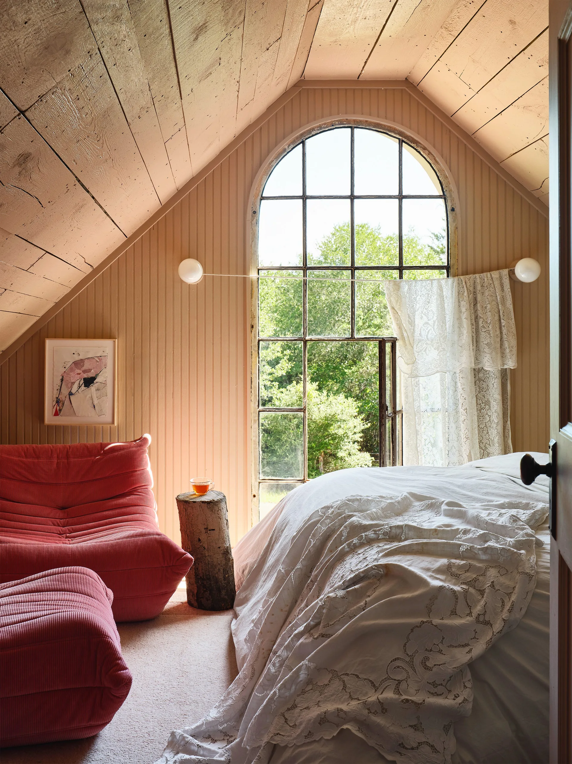 An attic bedroom featuring a large arched window with a lace curtain, a sloped wood-plank ceiling, white bedding, and a ribbed coral lounge chair.
