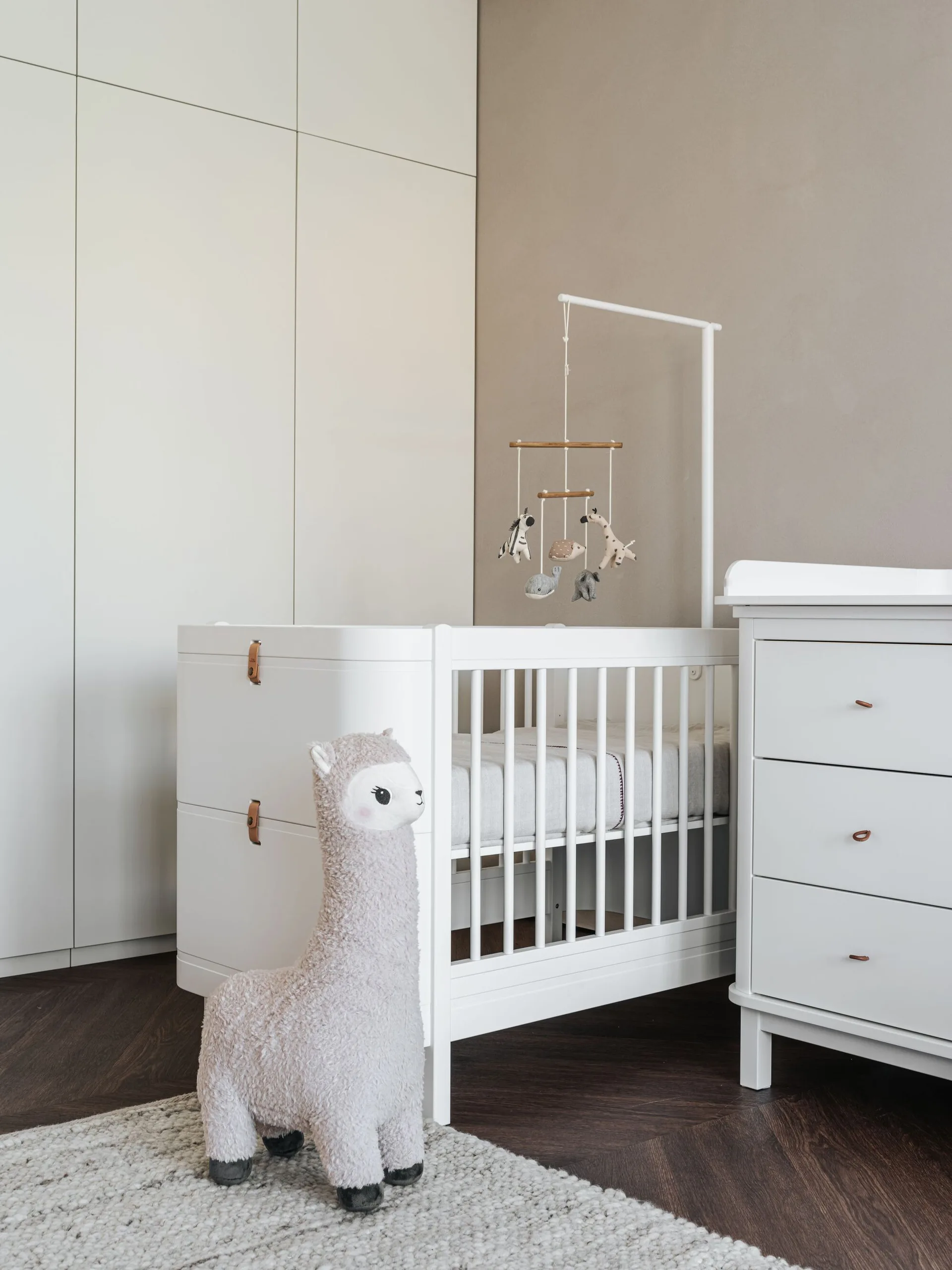 A white baby crib stands next to a matching dresser and built-in wardrobe in a neutral nursery with a plush alpaca toy on a textured rug.