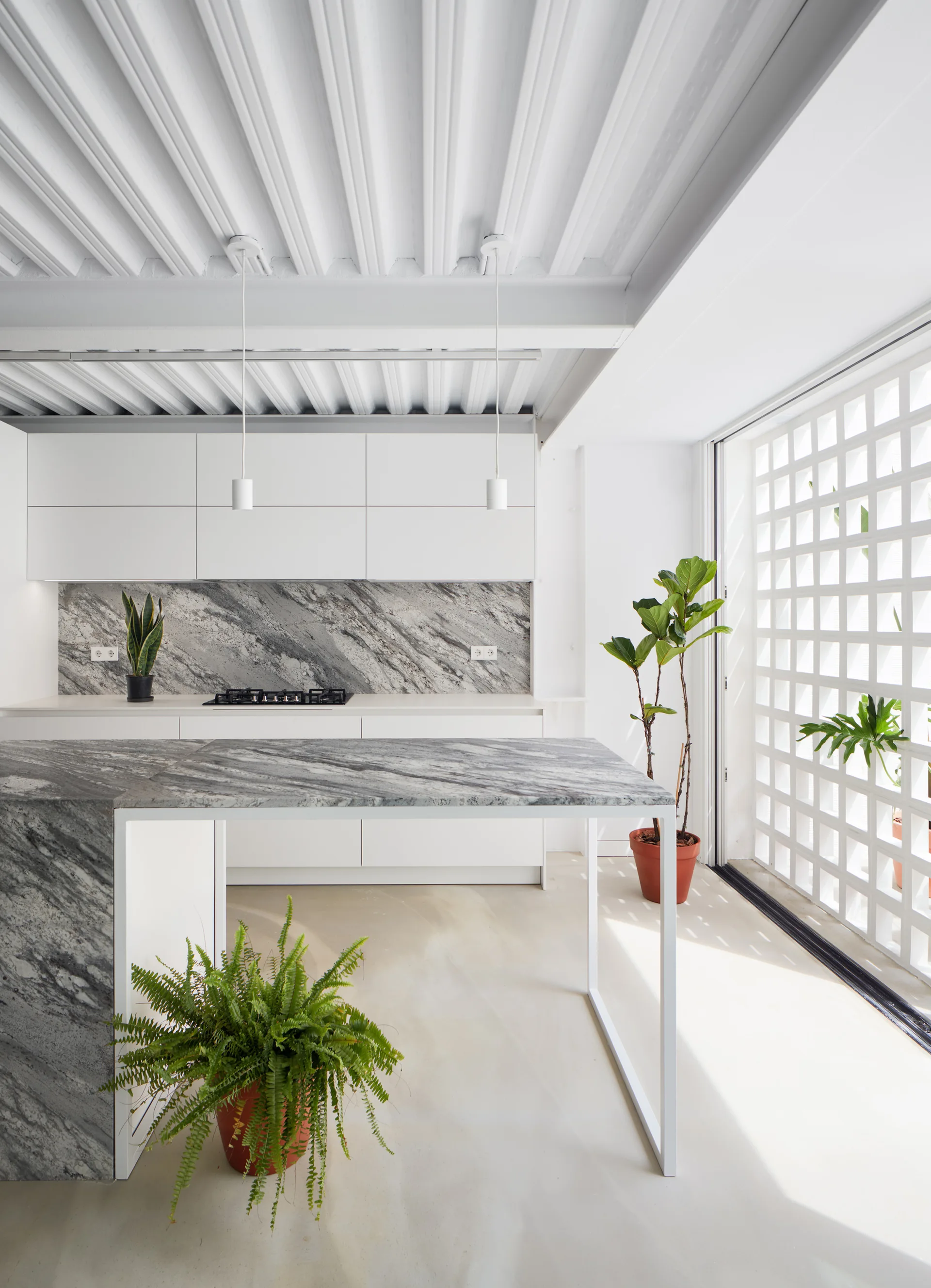 A minimalist white kitchen features a heavily veined grey marble island and backsplash, sleek white cabinetry, and a geometric breeze block wall partition.