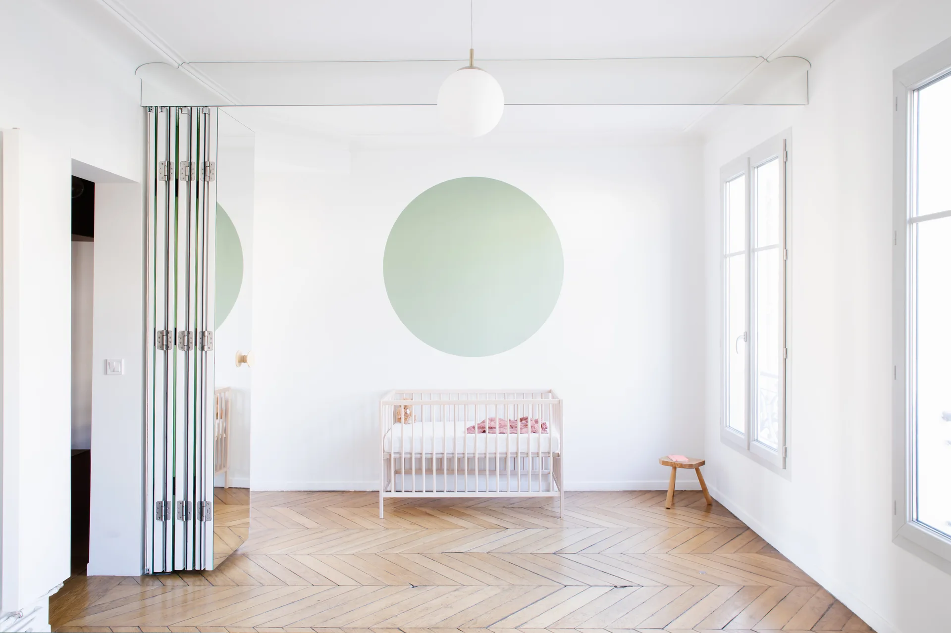 A bright nursery features a wooden crib in front of a white wall decorated with a large sage green circle and parquet herringbone flooring.