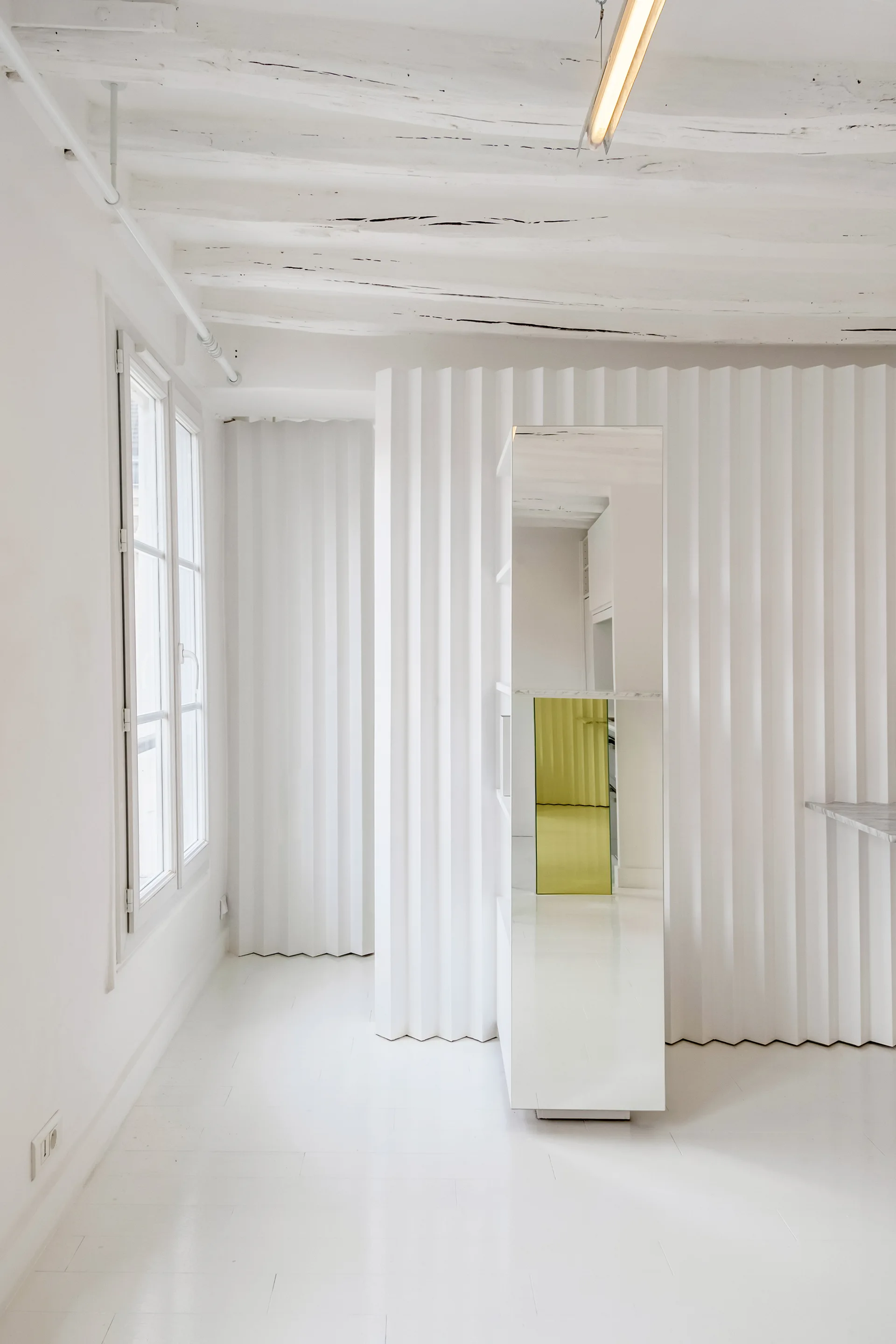 A bright white minimalist room featuring textured corrugated wall panels, exposed ceiling beams, a window, and a central freestanding mirrored cabinet.