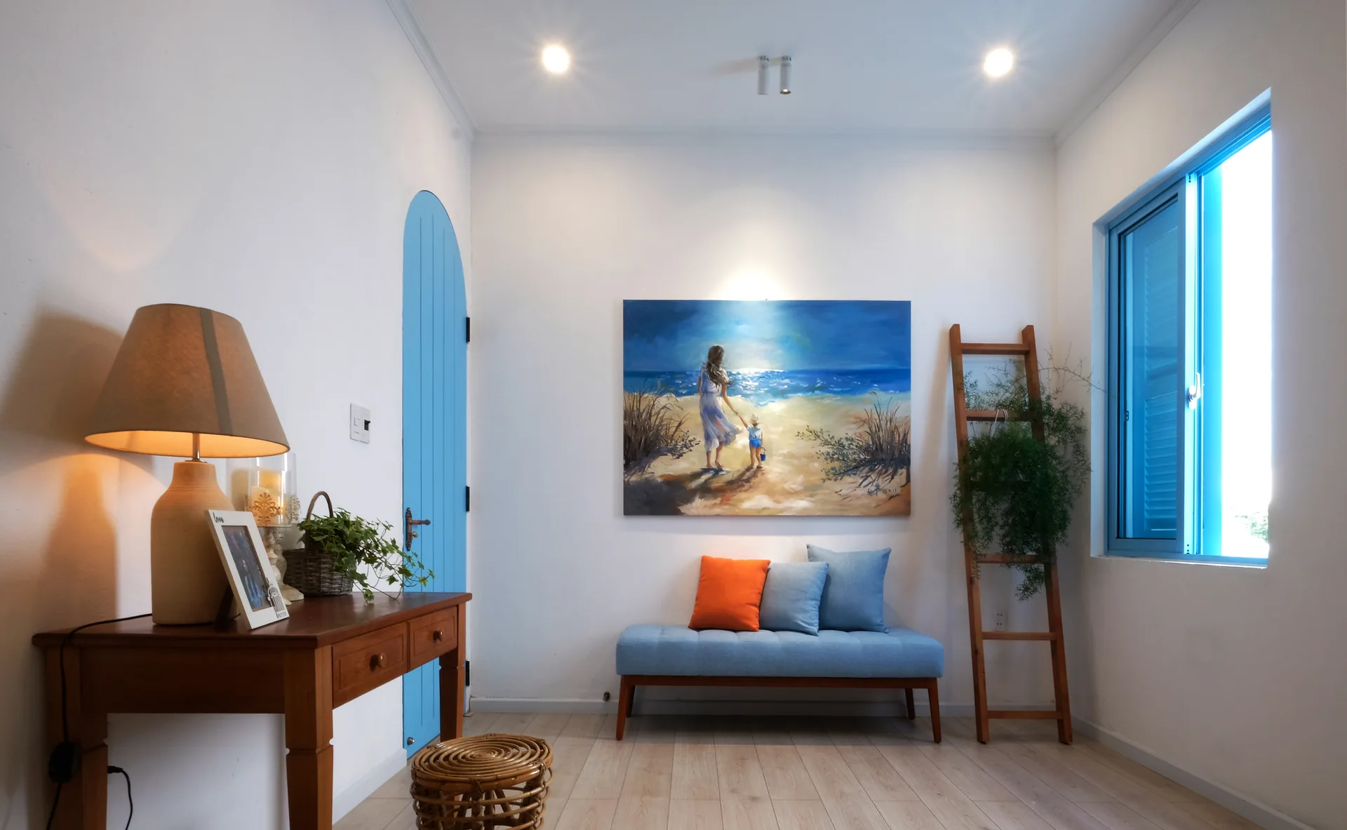 A bright entryway features a light wood floor, a blue upholstered bench under a beach painting, a wooden console table, and a blue arched door.