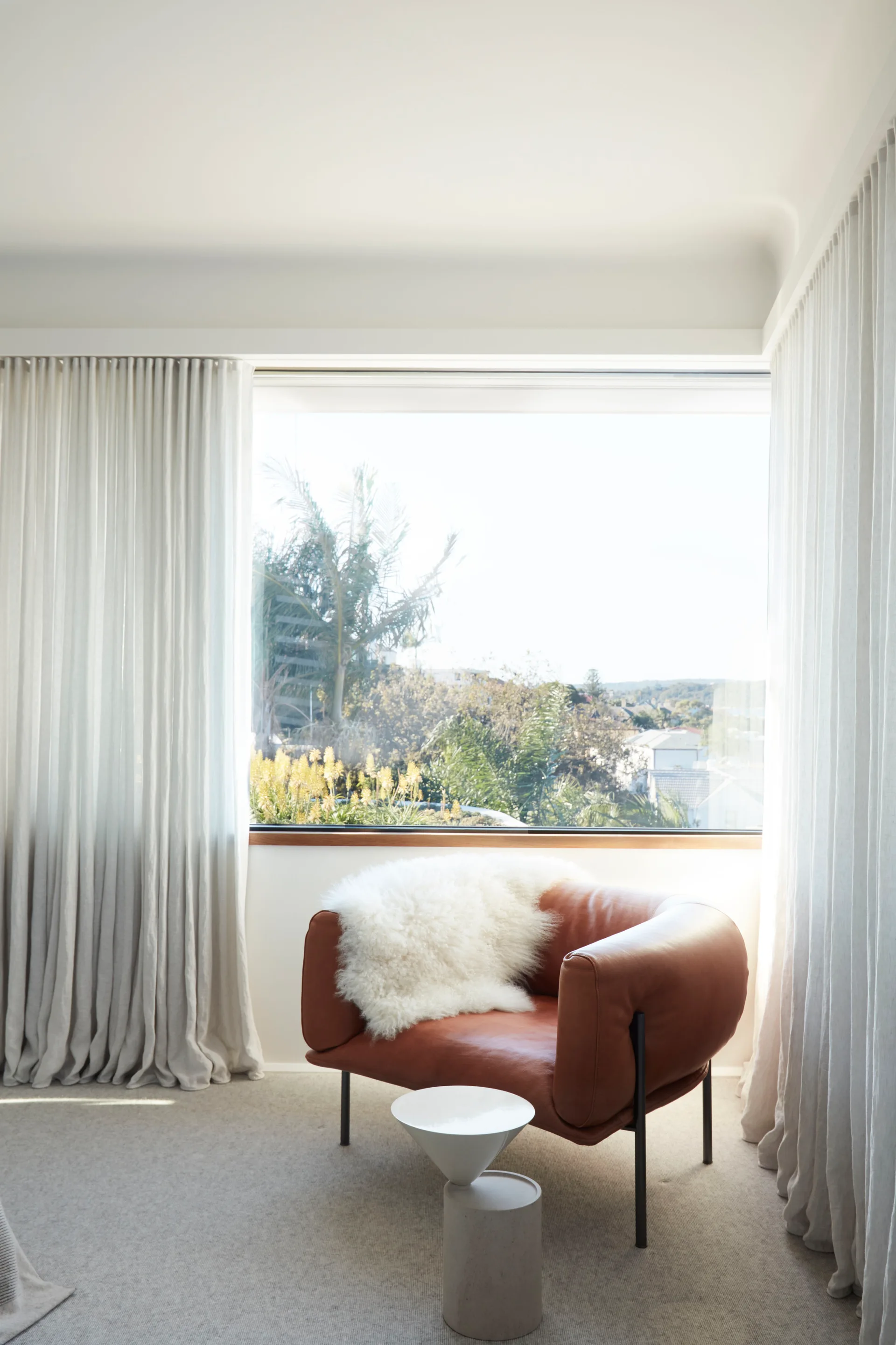 A cognac leather armchair draped with a white sheepskin throw sits by a large window with sheer floor-length curtains and a small white side table.