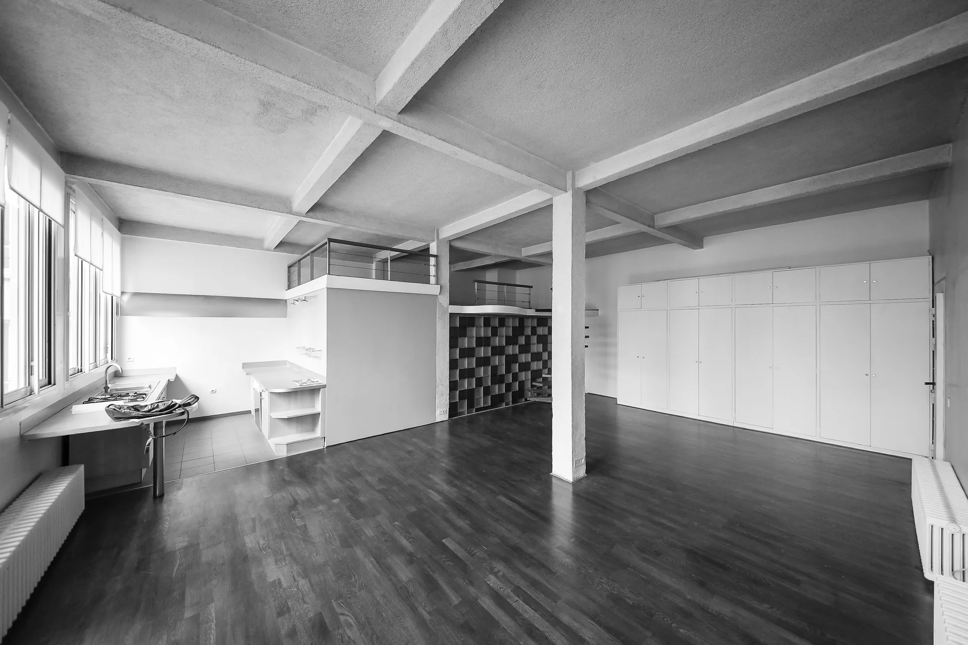 Spacious loft interior featuring an exposed concrete ceiling grid, dark wood floors, and a wall of white built-in storage cabinets.