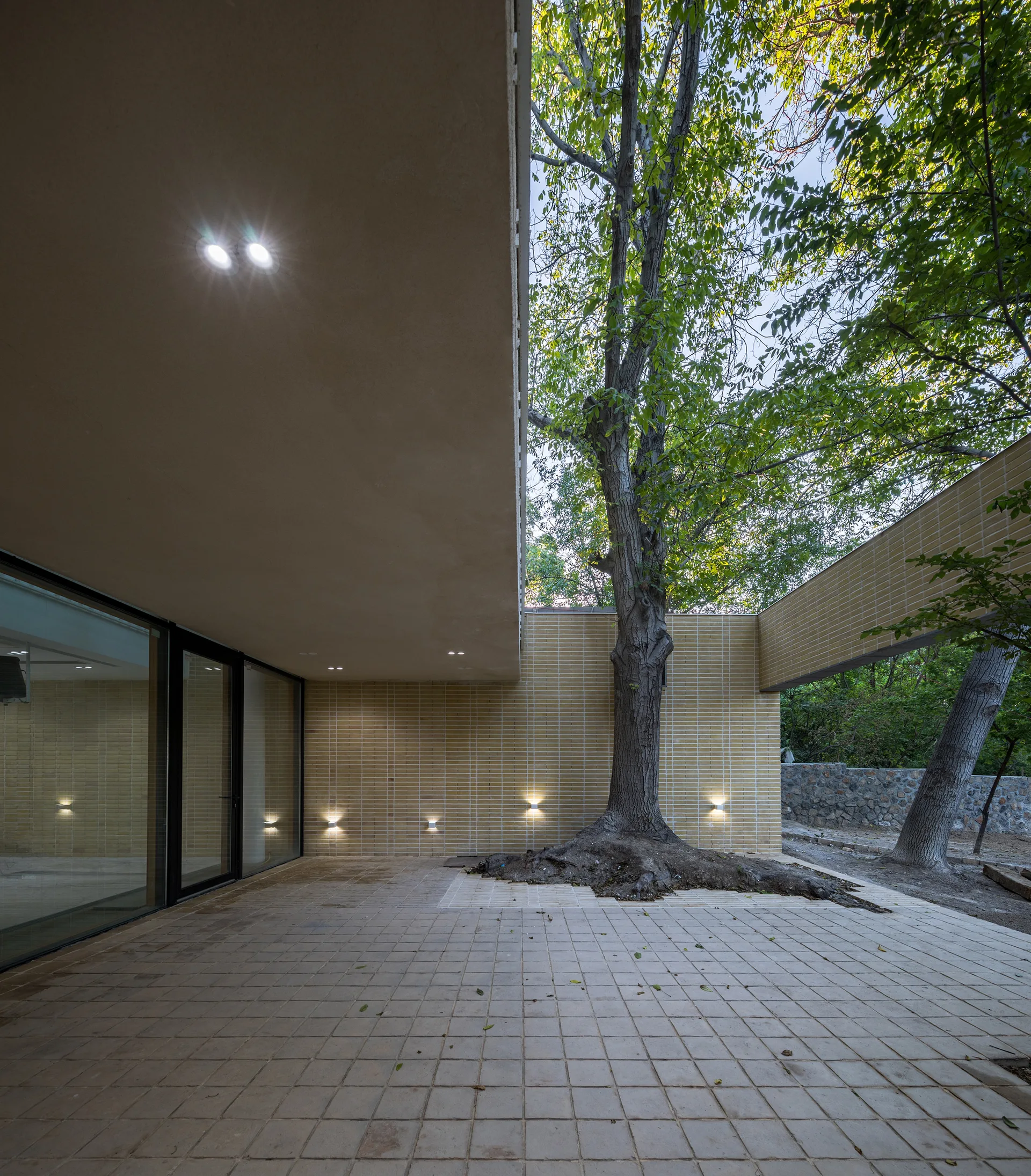 A covered modern patio with a large tree growing through the structure, light tan brick walls, wall sconces, and square paved floors.