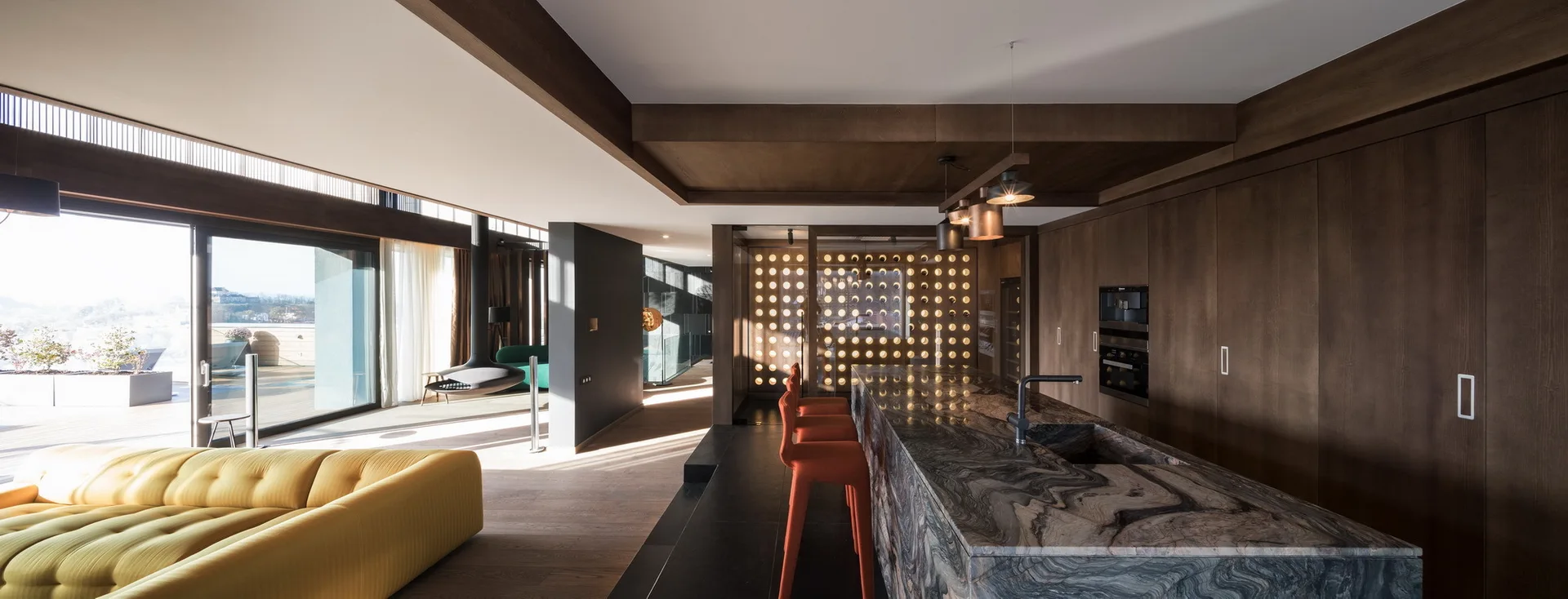 An open-concept kitchen features a large veined marble island, orange bar stools, dark wood cabinets, and an illuminated circular patterned screen.