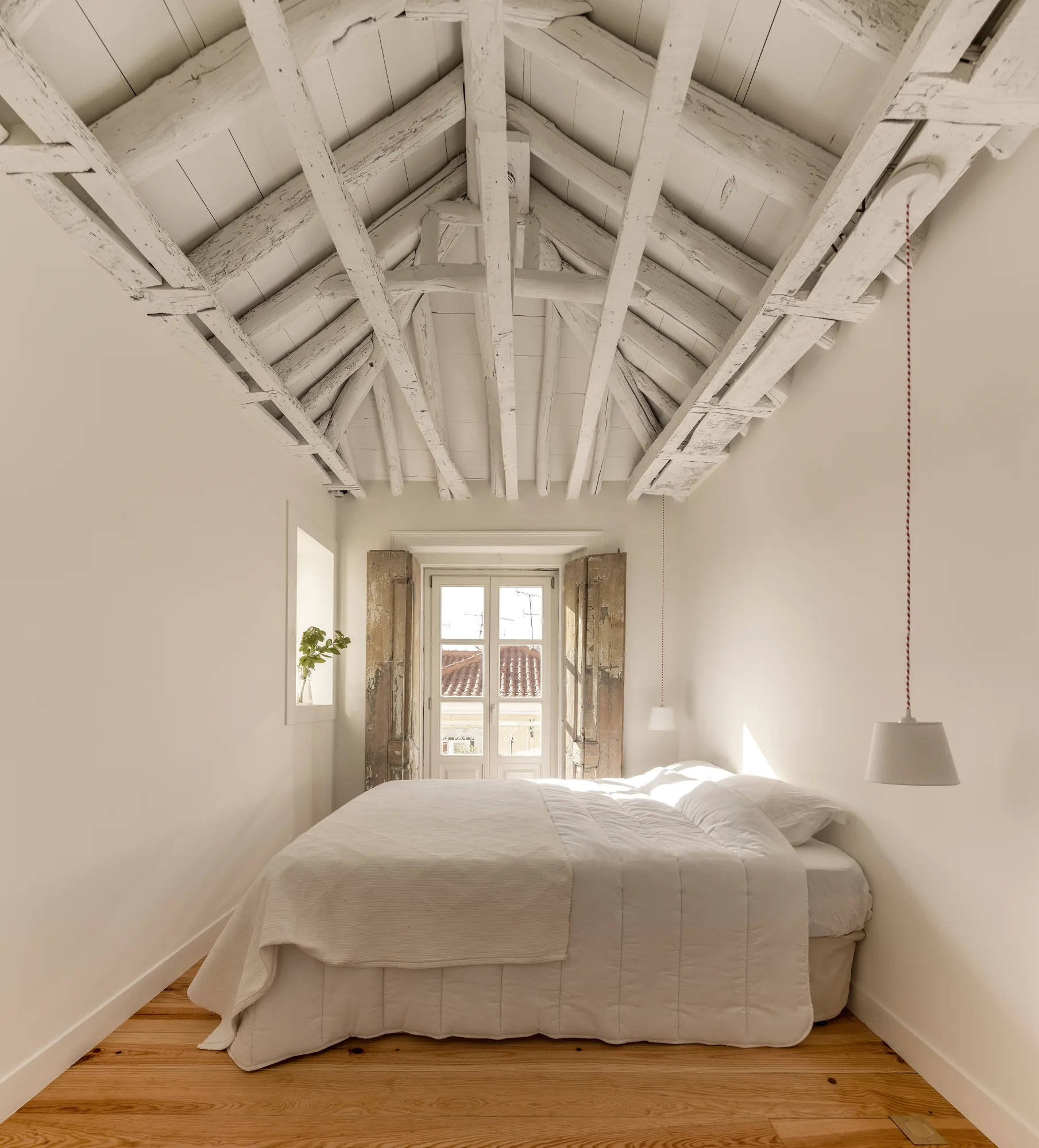 A white bedroom with a bed centered under a vaulted ceiling with exposed white beams, featuring distressed wooden shutters on the window.