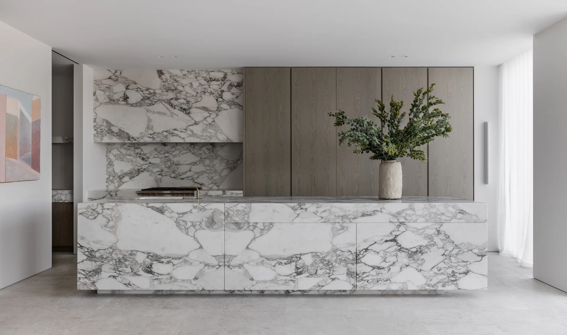 A minimalist kitchen features a large veined white marble island, matching marble backsplash, and seamless vertical wood cabinetry.