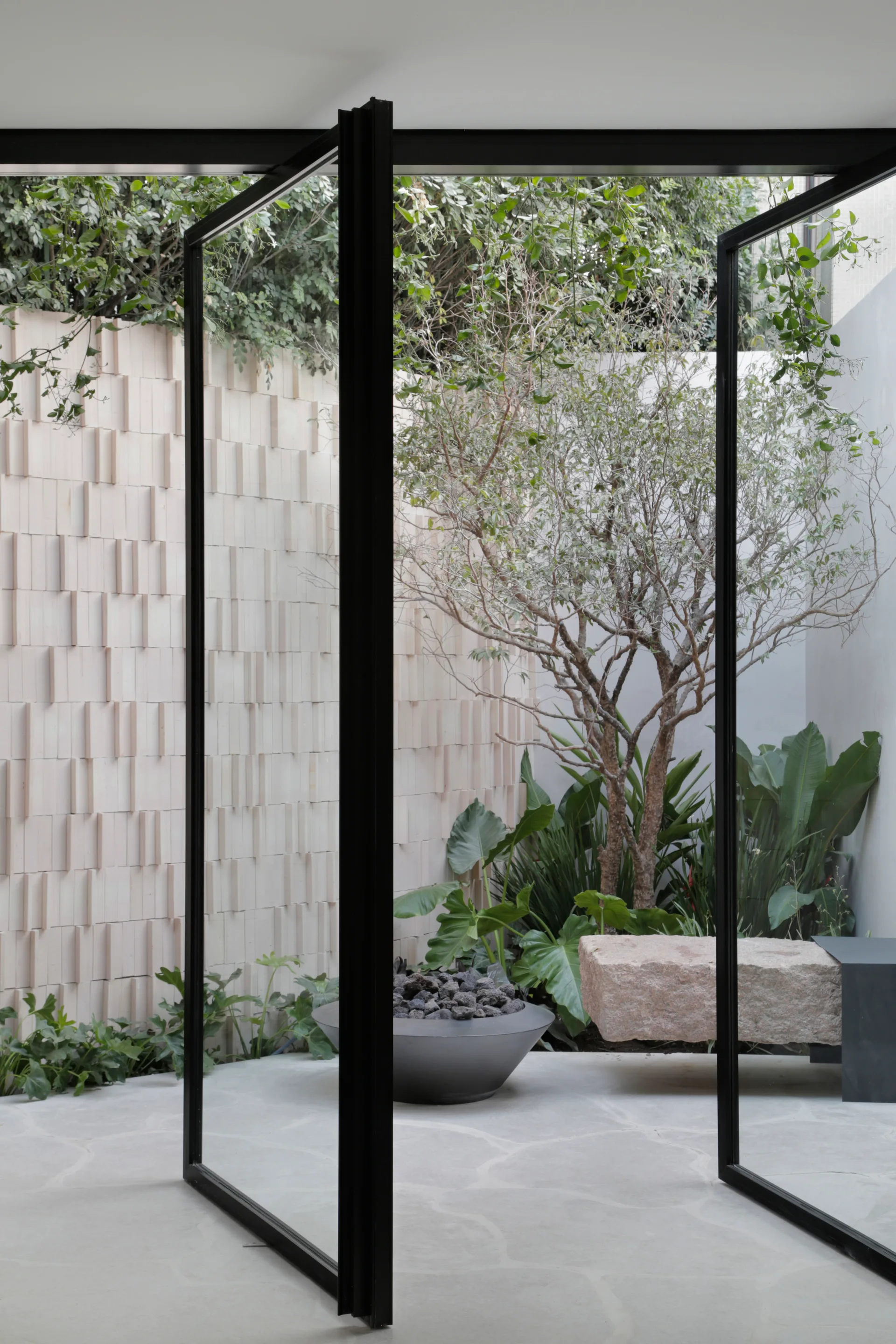 A view through black-framed glass pivot doors into a minimalist courtyard with a staggered brick wall, stone bench, and green plants.